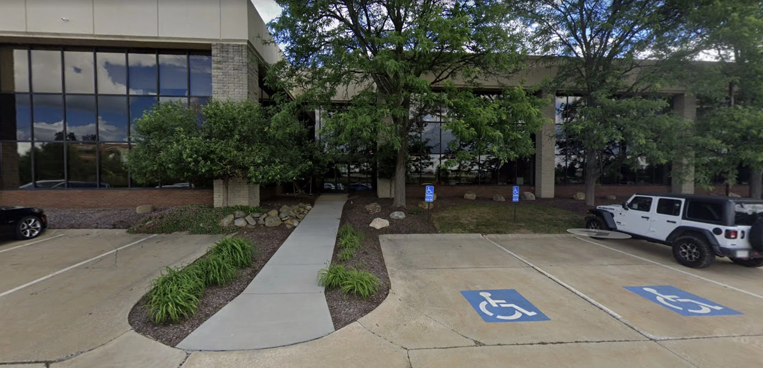 Sidewalk leading up to a two-door entrance with tress on both sides. The two buildings with windows are visible on the left and right. Two accessible parking spaces are in the foreground.