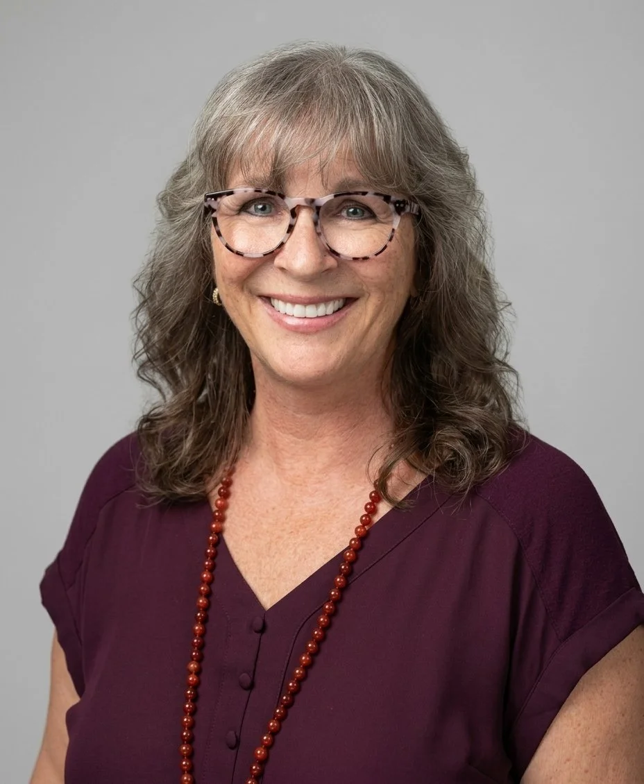 A professional 3/4 portrait headshot of Vickie with wavy gray-brown hair, bangs, tortoiseshell glasses, and a warm smile. She is wearing a deep purple short-sleeved blouse and a long red beaded necklace set against a plain, soft grey studio backdrop.