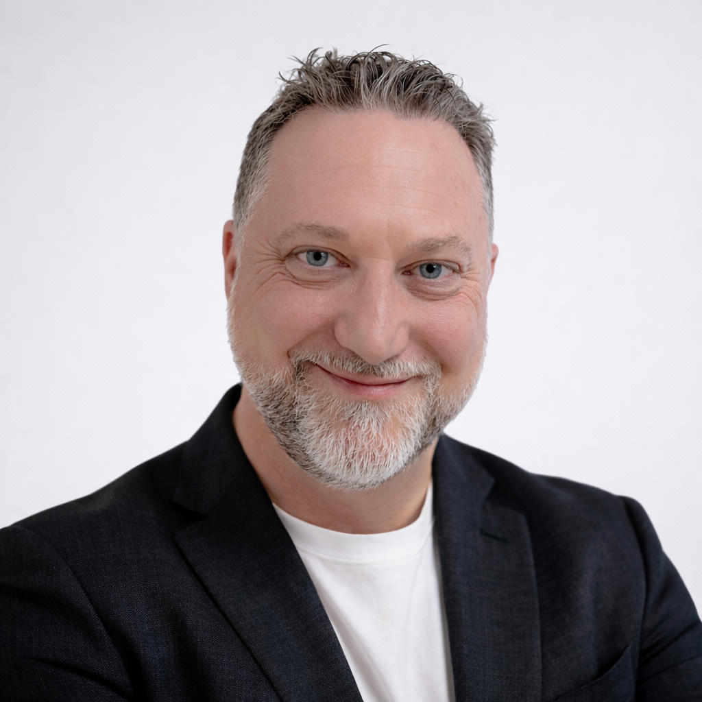 A middle-aged man with gray hair and beard, smiling, wearing a black blazer and white t-shirt against a plain white background.