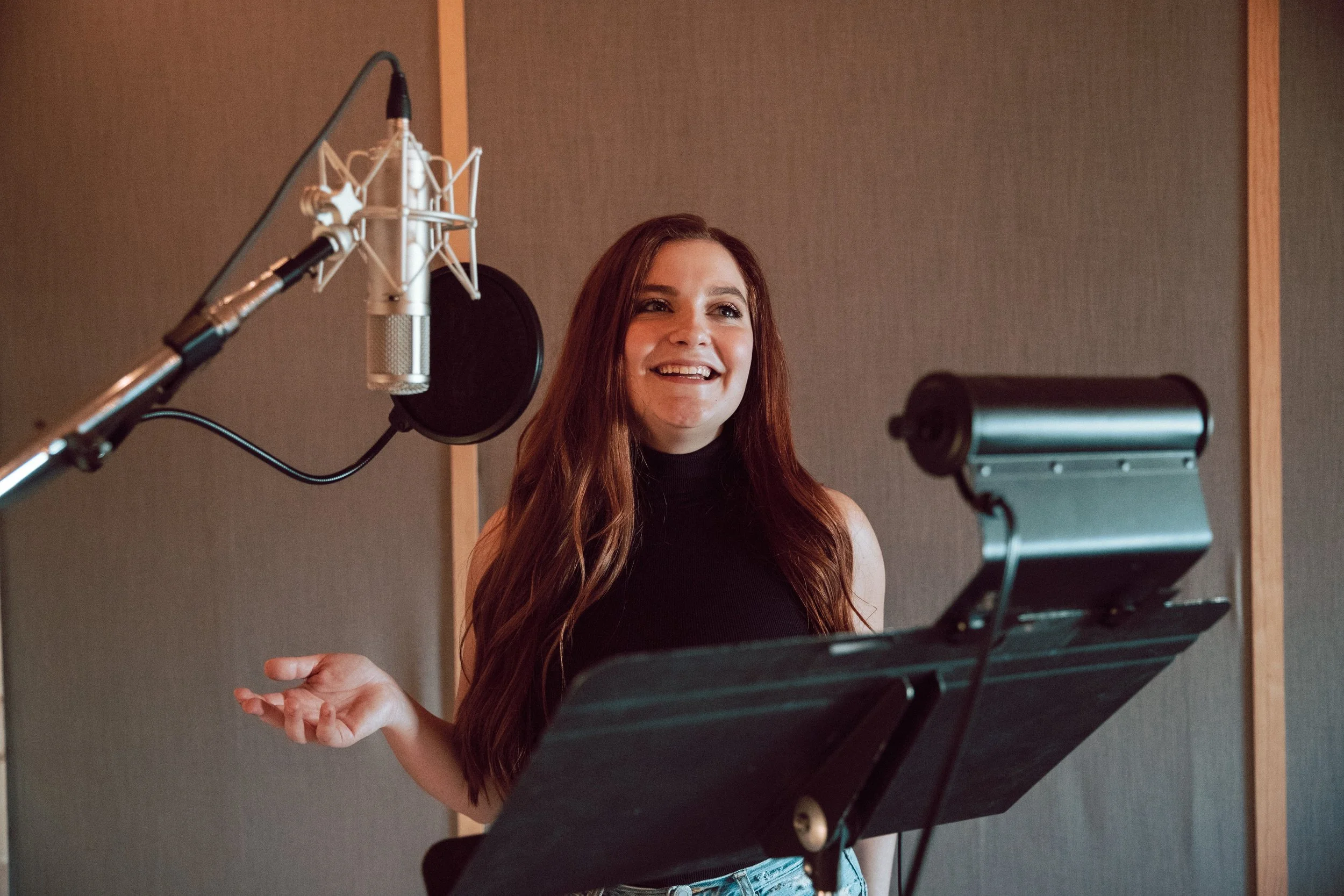 A young woman with long red hair wearing a black sleeveless turtleneck, standing in a recording studio with a microphone and music stand, smiling and gesturing with her hand.