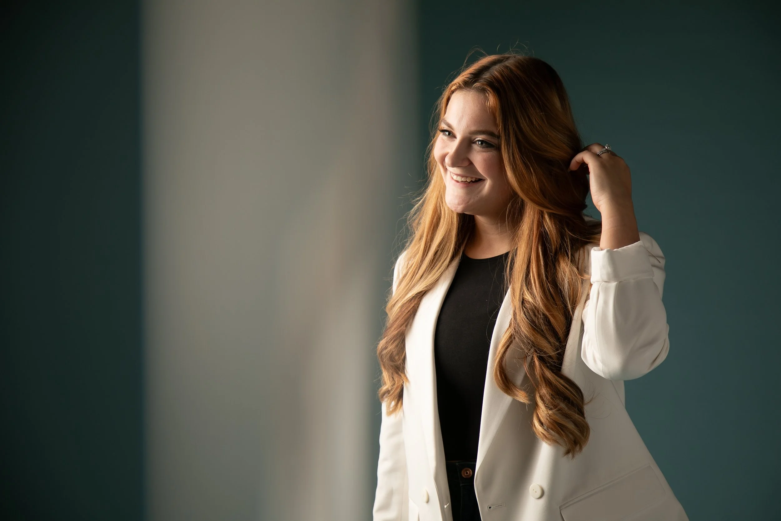 Smiling woman with long, wavy red hair wearing a white blazer and a black top, standing in front of a teal-colored wall.