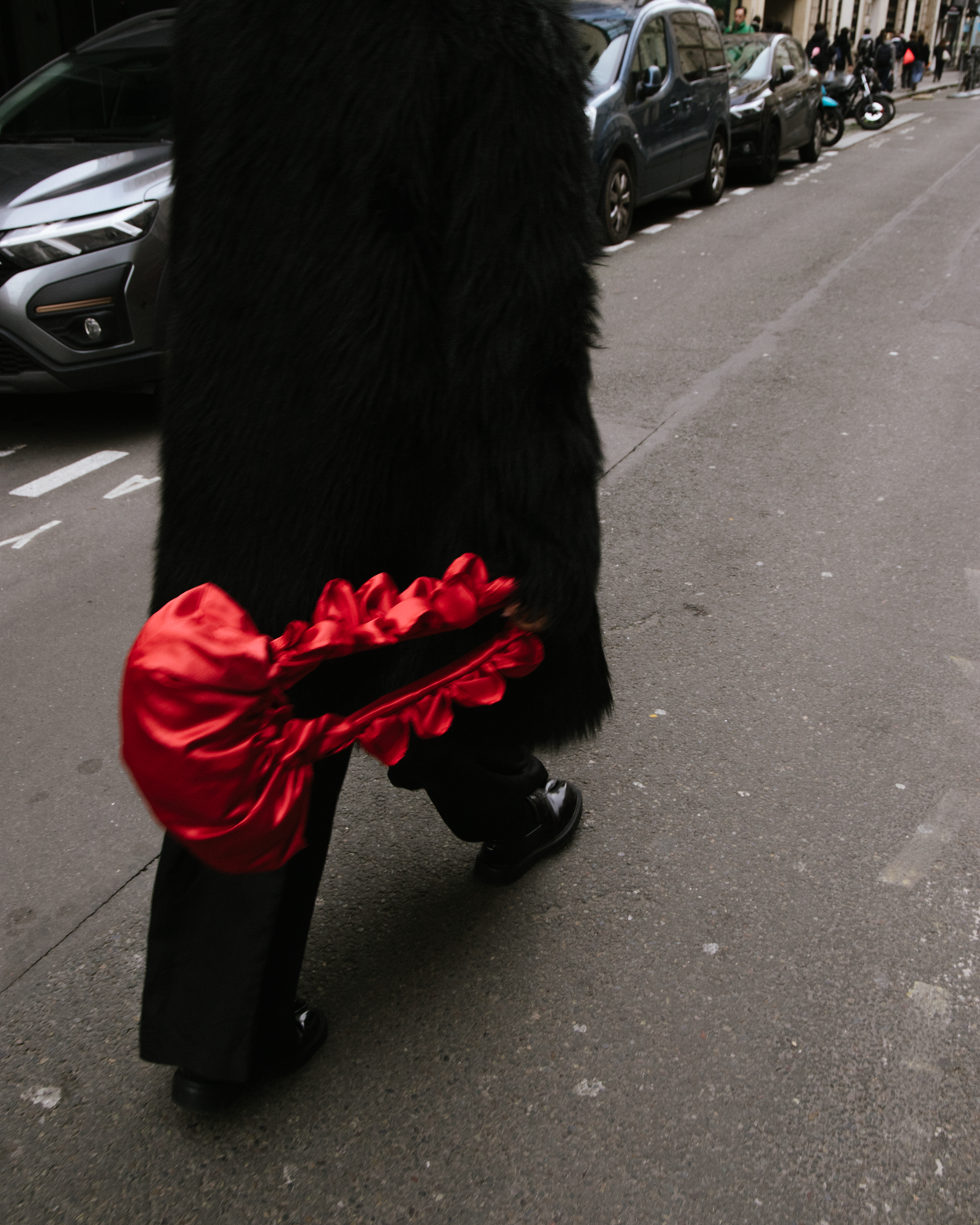 Person wearing a black fur coat and black shoes walking on a city sidewalk, carrying a shiny red satin bag with ruffled edges.
