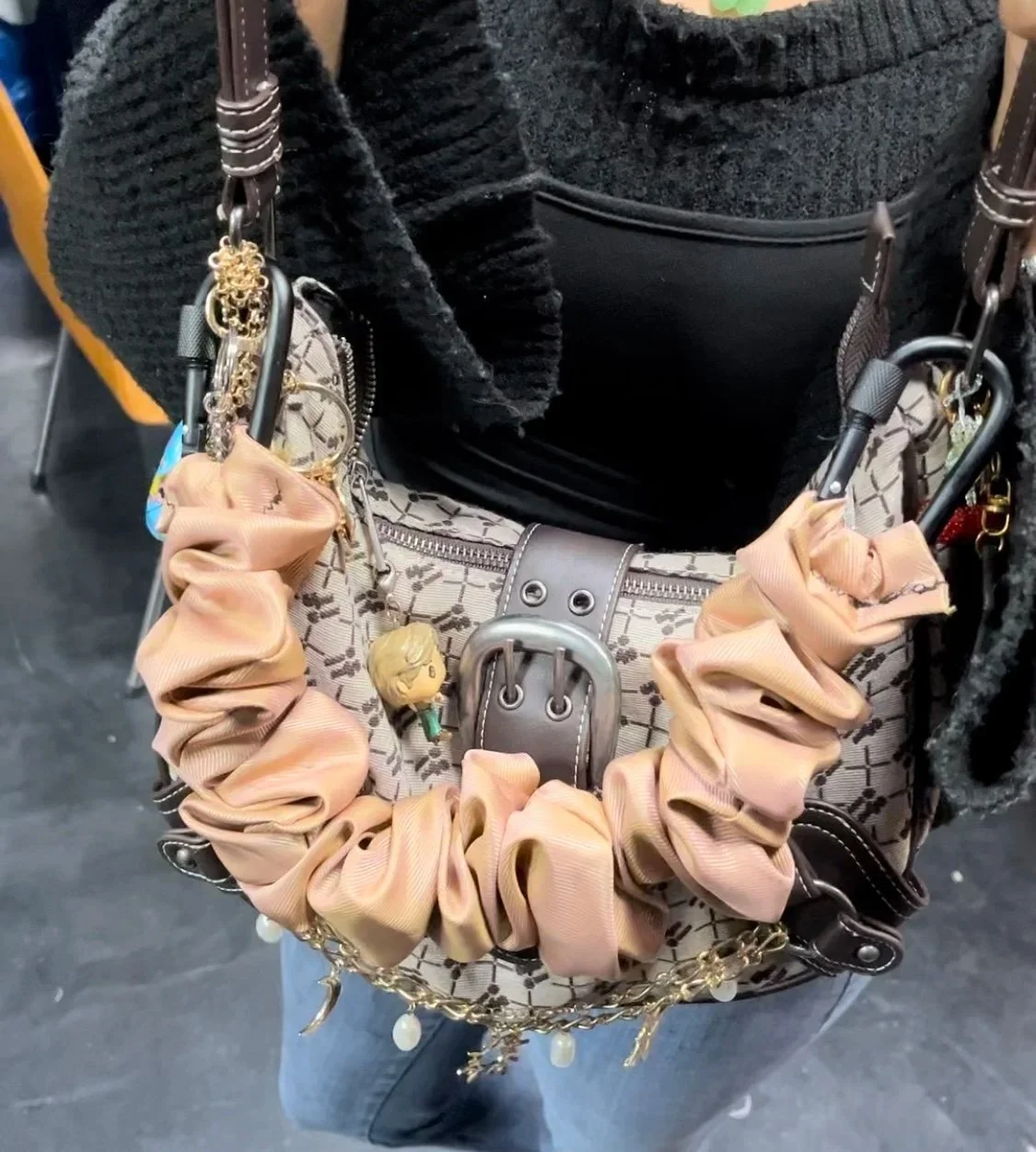 A purse decorated with a pink scrunchie and charm, hanging on a person holding a black and beige patterned bag with a black zippered pocket and silver buckle.
