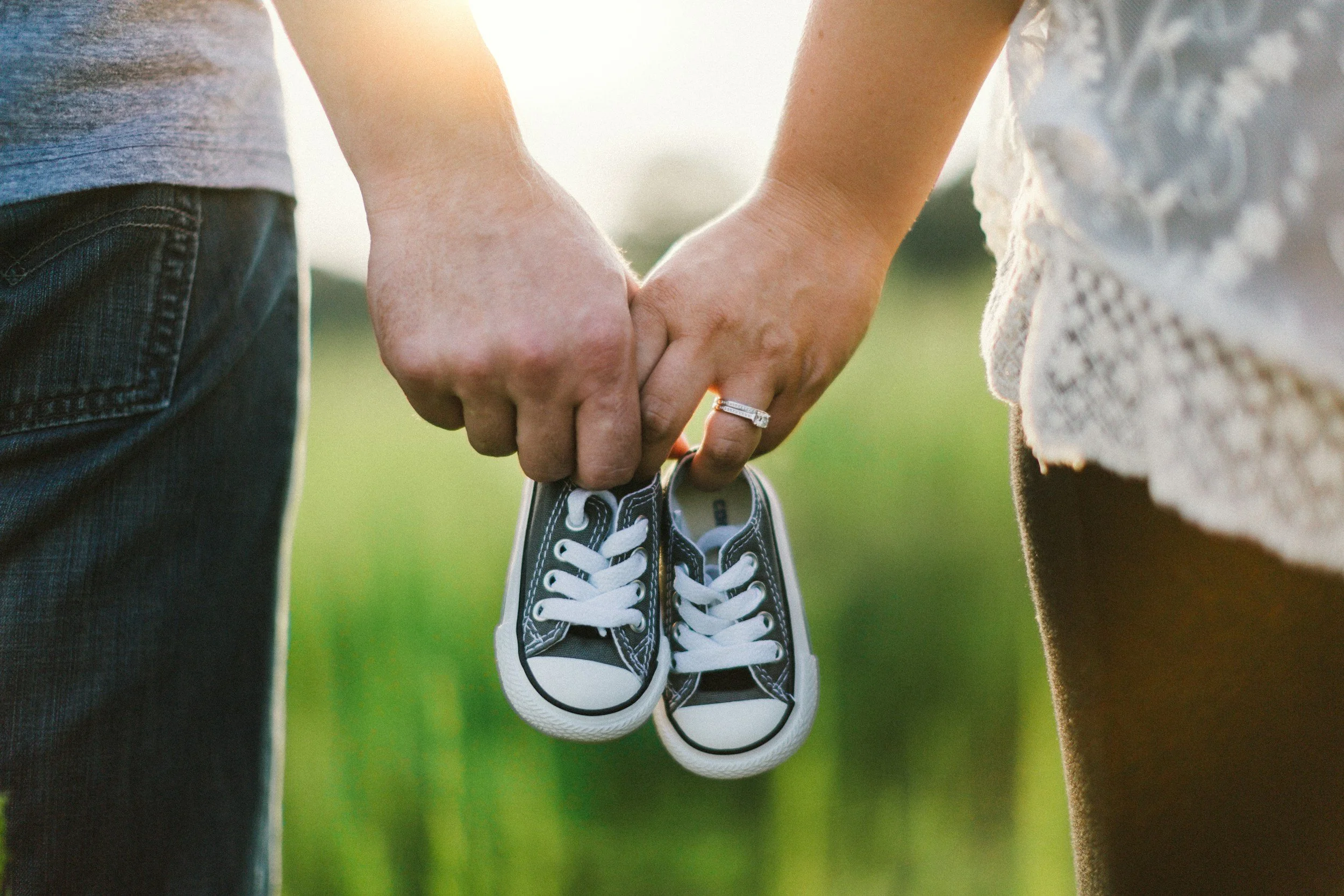 Close-up of two people holding hands, with a pair of small black and white baby sneakers hanging from their fingers . The background is a blurred outdoor setting with sunlight.