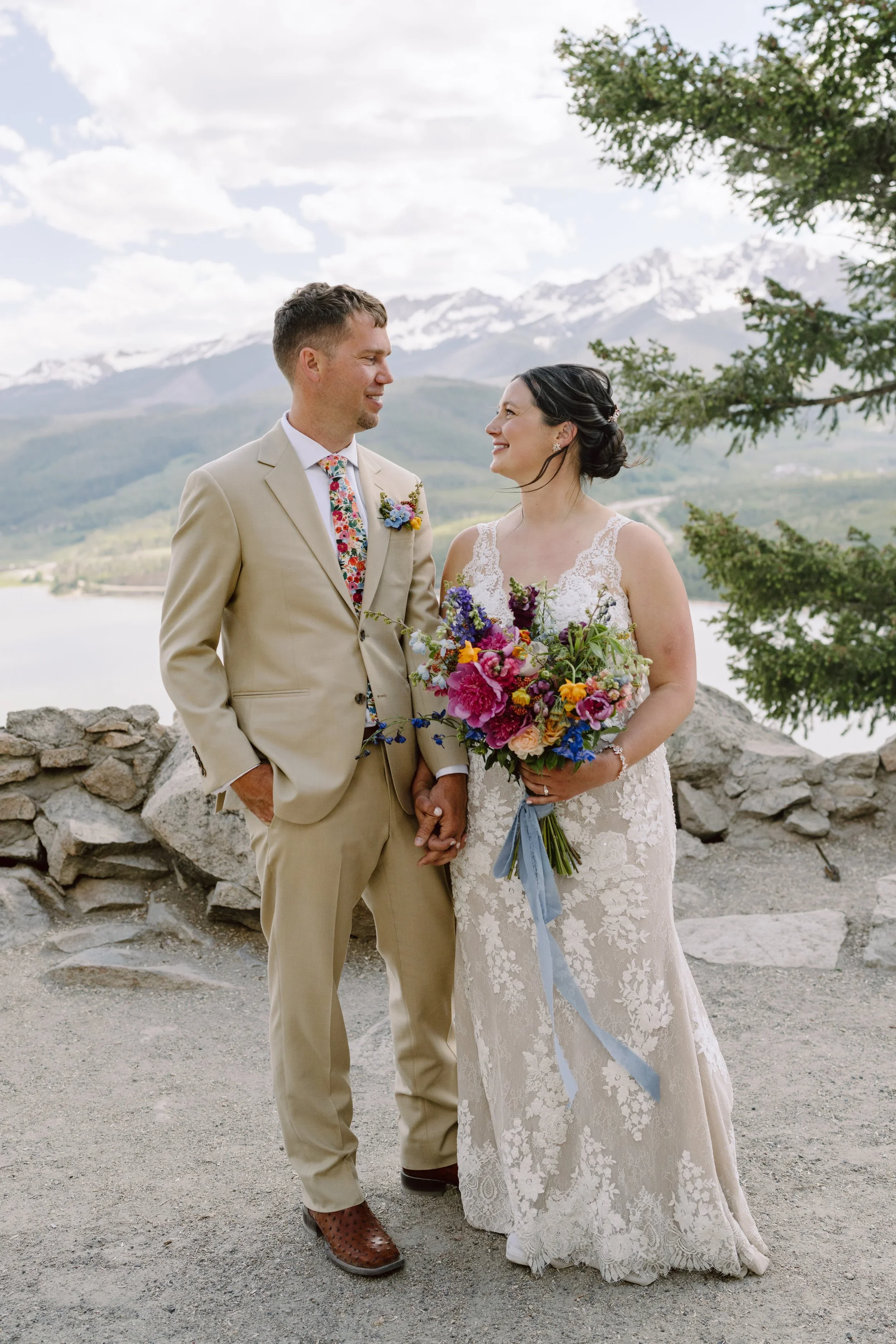 A couple dressed in wedding attire holding hands outdoors with mountains and a lake in the background, woman holding a colorful bouquet.