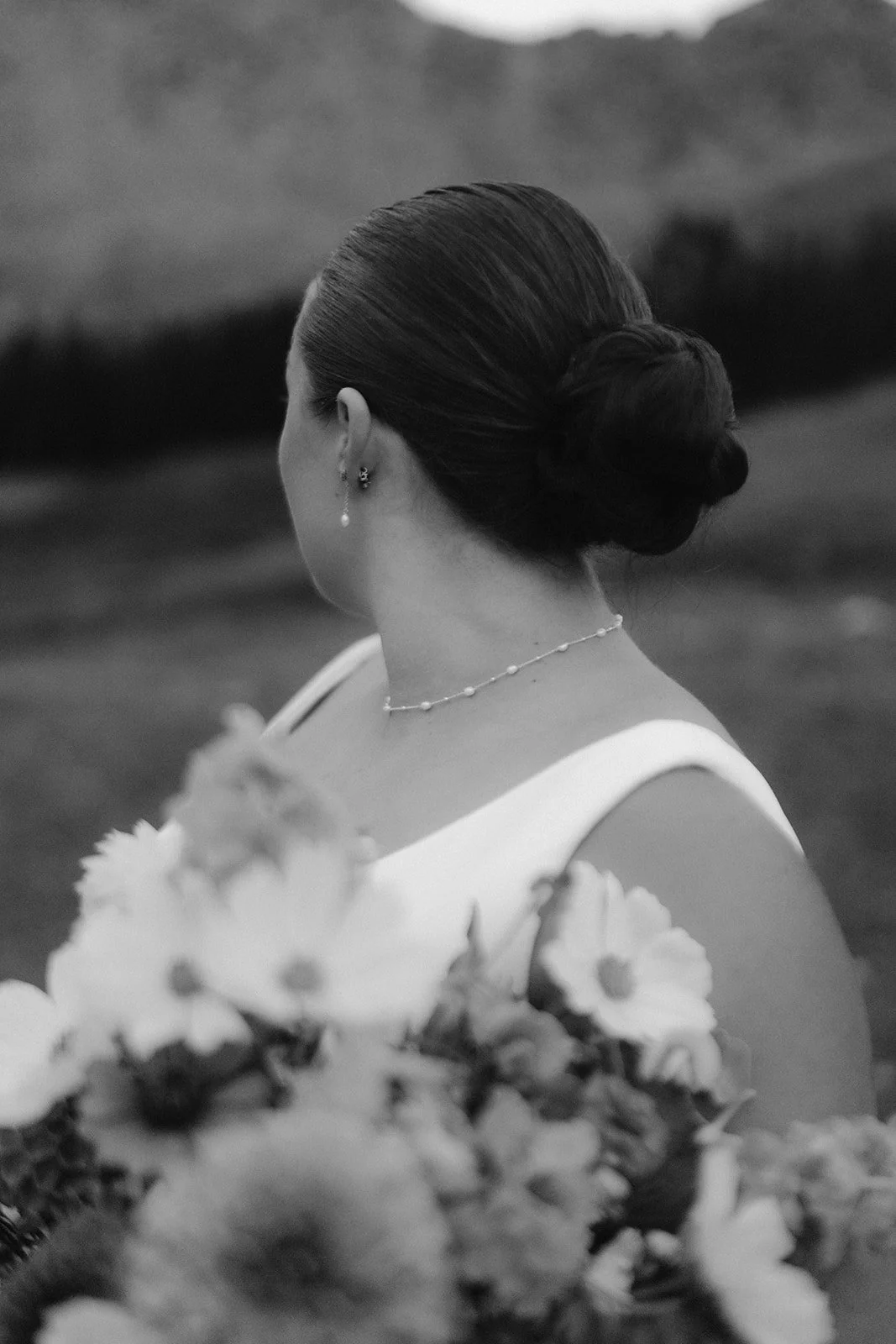 A woman with her hair styled in an elegant updo, wearing earrings and a necklace, holding a bouquet of flowers, with blurred outdoor landscape in the background.