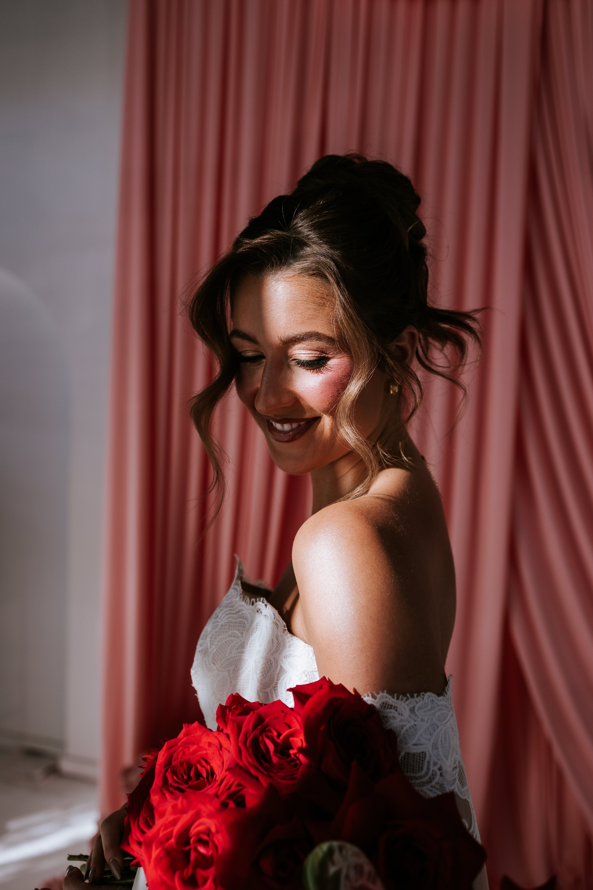 A woman in a white lace dress holding a bouquet of red roses, smiling with her eyes closed, standing in front of pink curtains.
