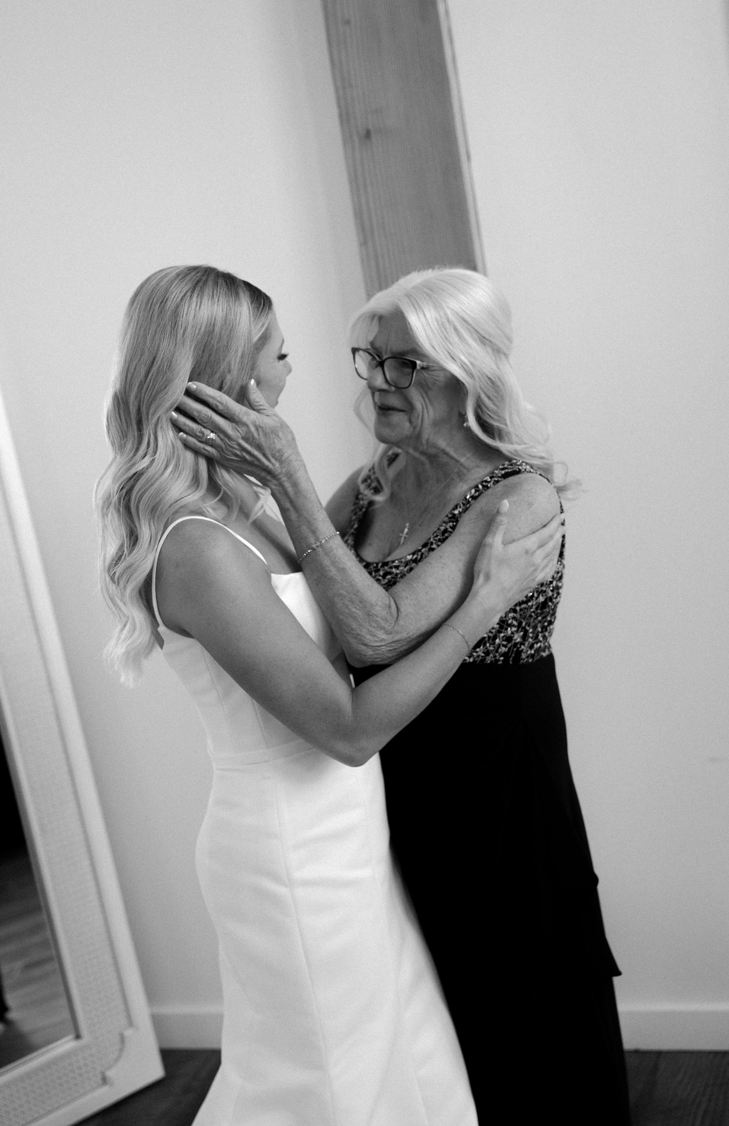 An elderly woman with glasses embraces a young woman in a wedding dress, holding her face tenderly in a heartfelt moment.