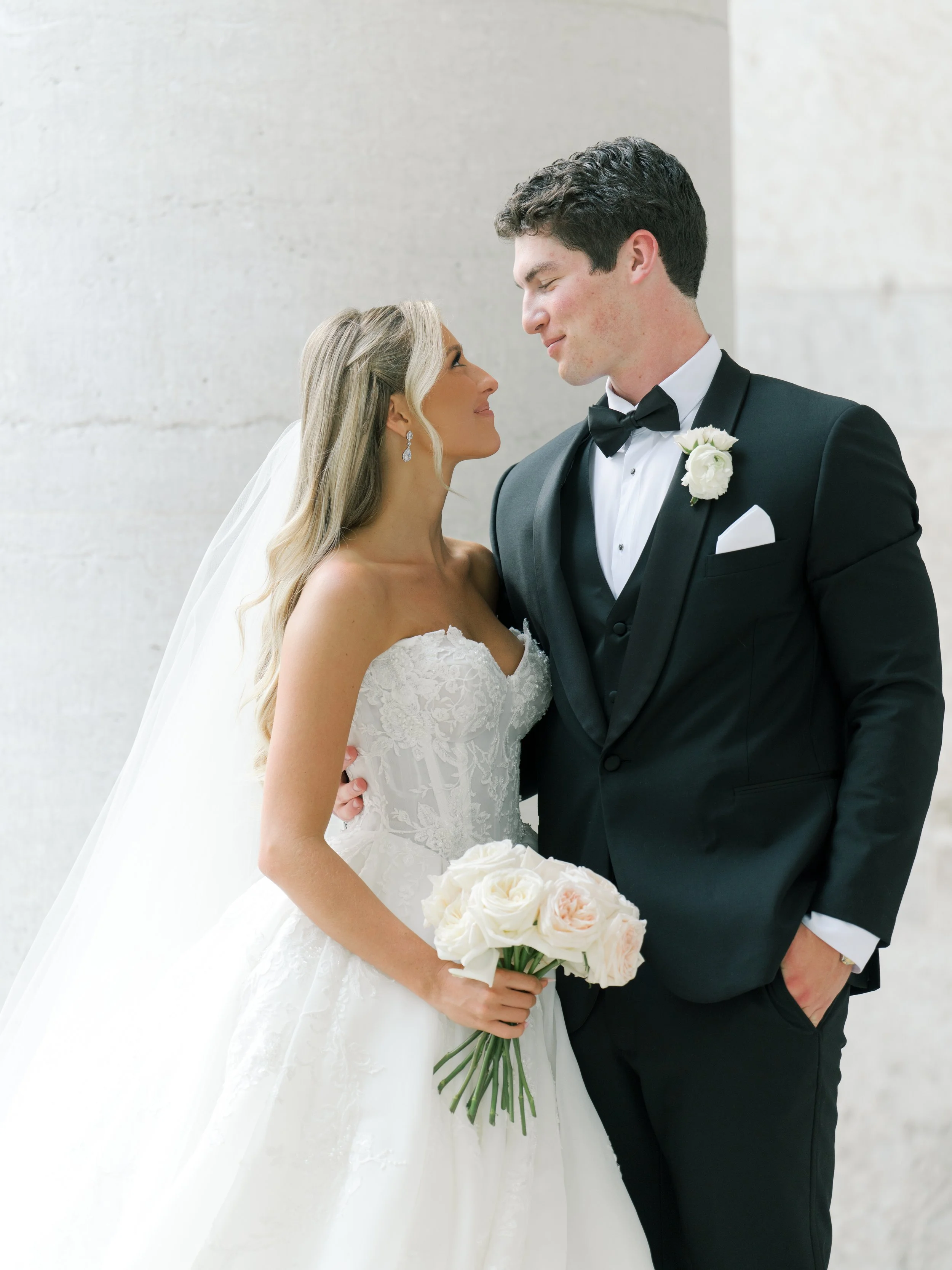 Bride and groom standing close, looking into each other's eyes, smiling. The bride is wearing a white lace wedding gown, holding a bouquet of white roses, and has long blonde hair with a veil. The groom is dressed in a black tuxedo with a white shirt, black bow tie, and a white boutonniere.