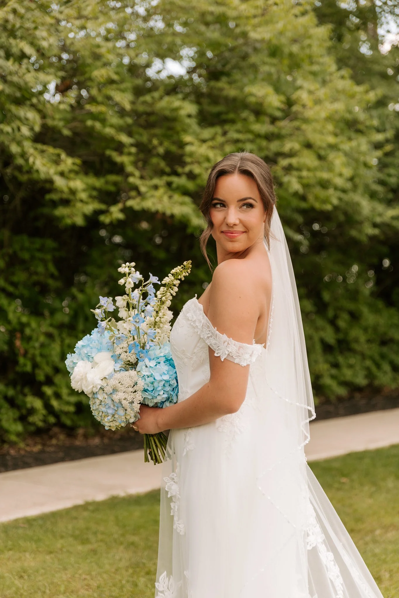 A bride in a white wedding dress holding a bouquet of blue and white flowers outdoors with greenery in the background.