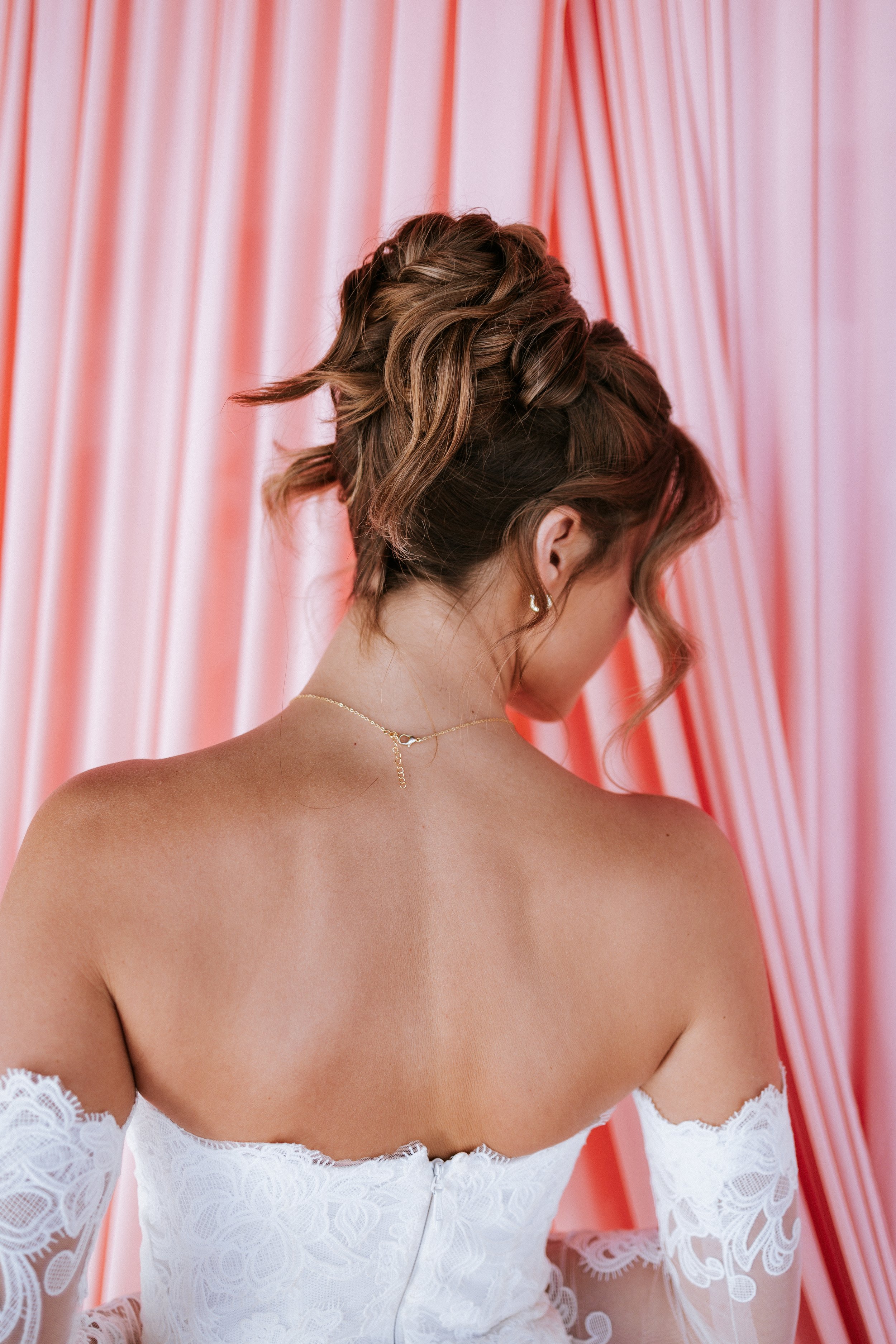 Bride with an elegant updo hairstyle, wearing jewelry, and a white lace off-shoulder wedding dress, standing in front of pink and red curtains.