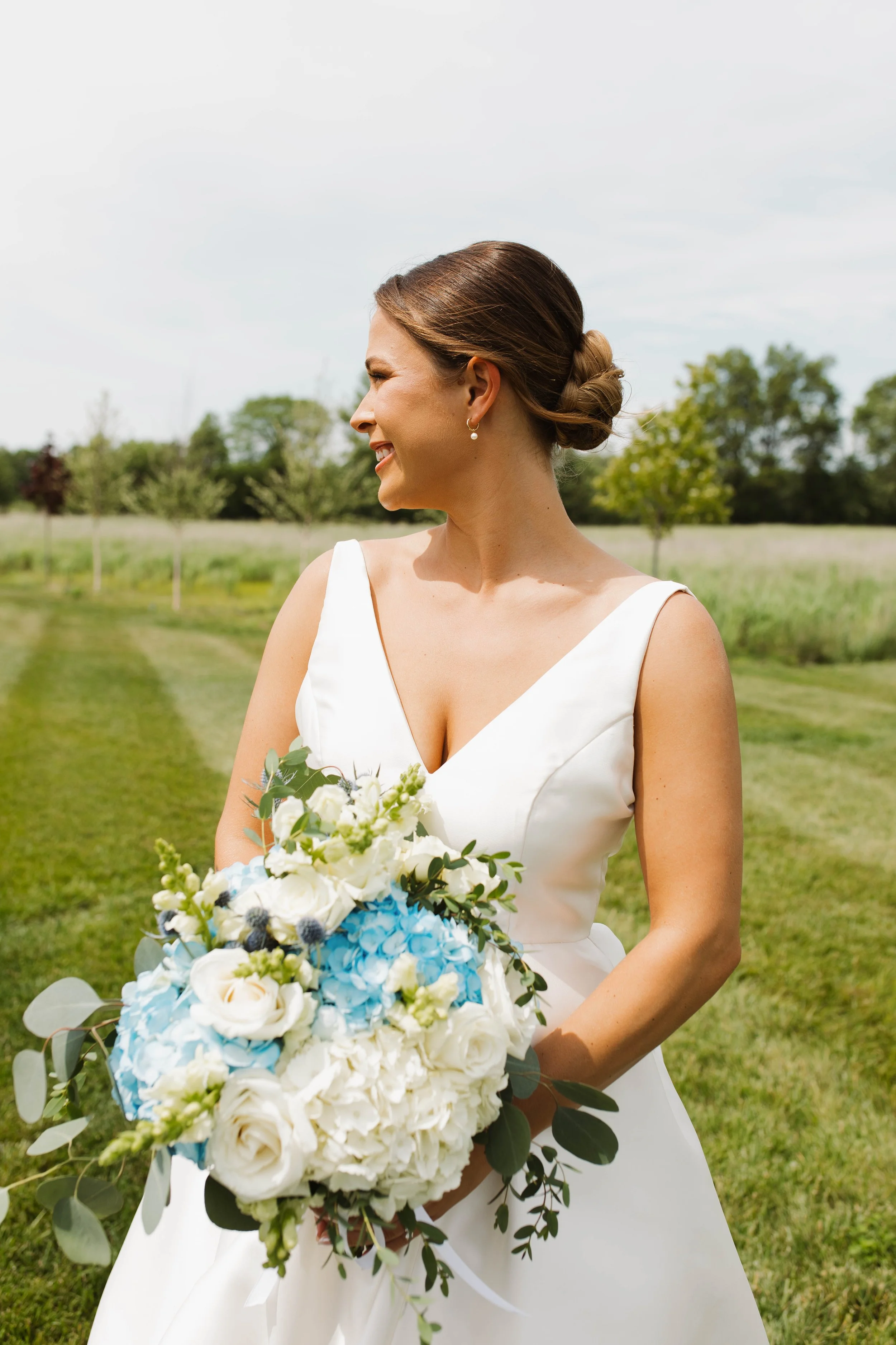 A bride in a white wedding dress holding a bouquet of flowers outdoors on a sunny day, smiling and looking to her left.