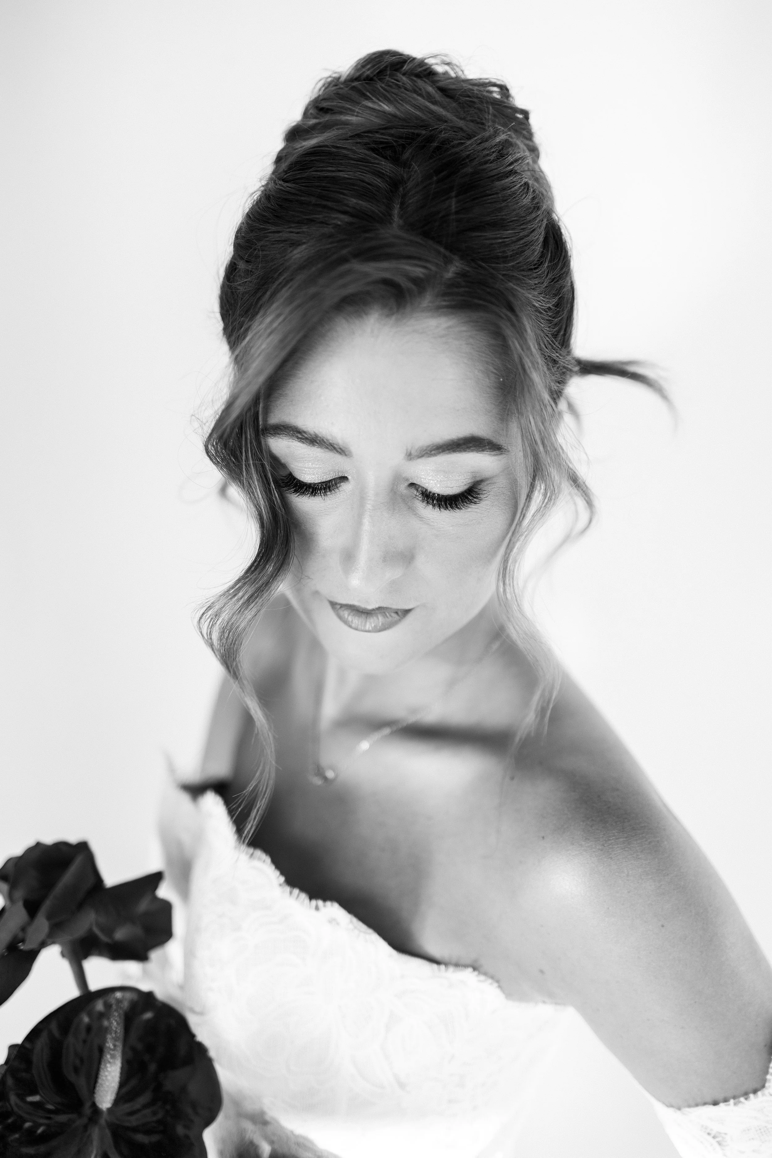Black and white portrait of a woman with styled hair, long lashes, and makeup, wearing a strapless dress with lace details, holding a flower.