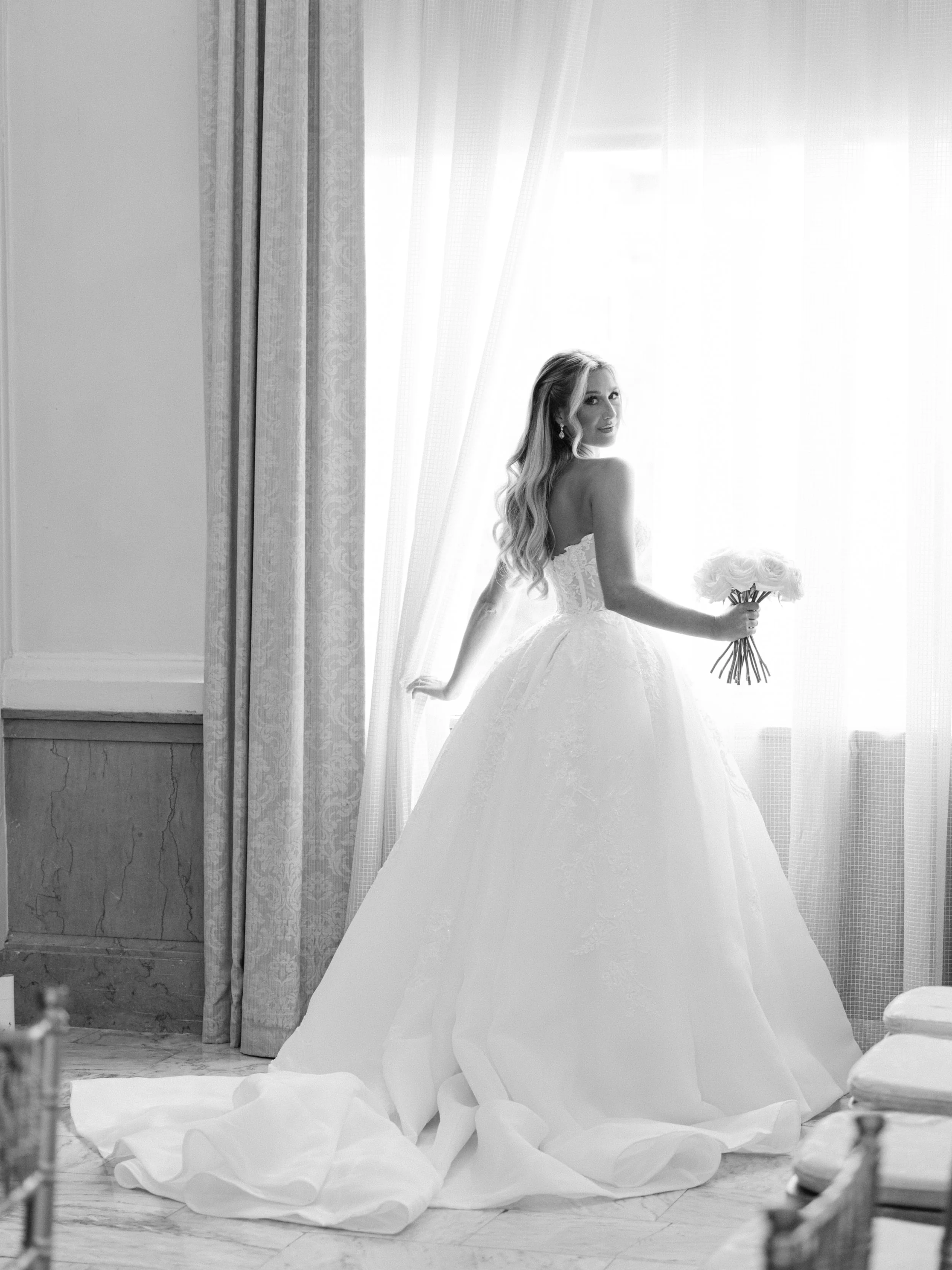 A bride in a wedding gown standing by a window, holding a bouquet of roses, with sunlight streaming through the curtains.