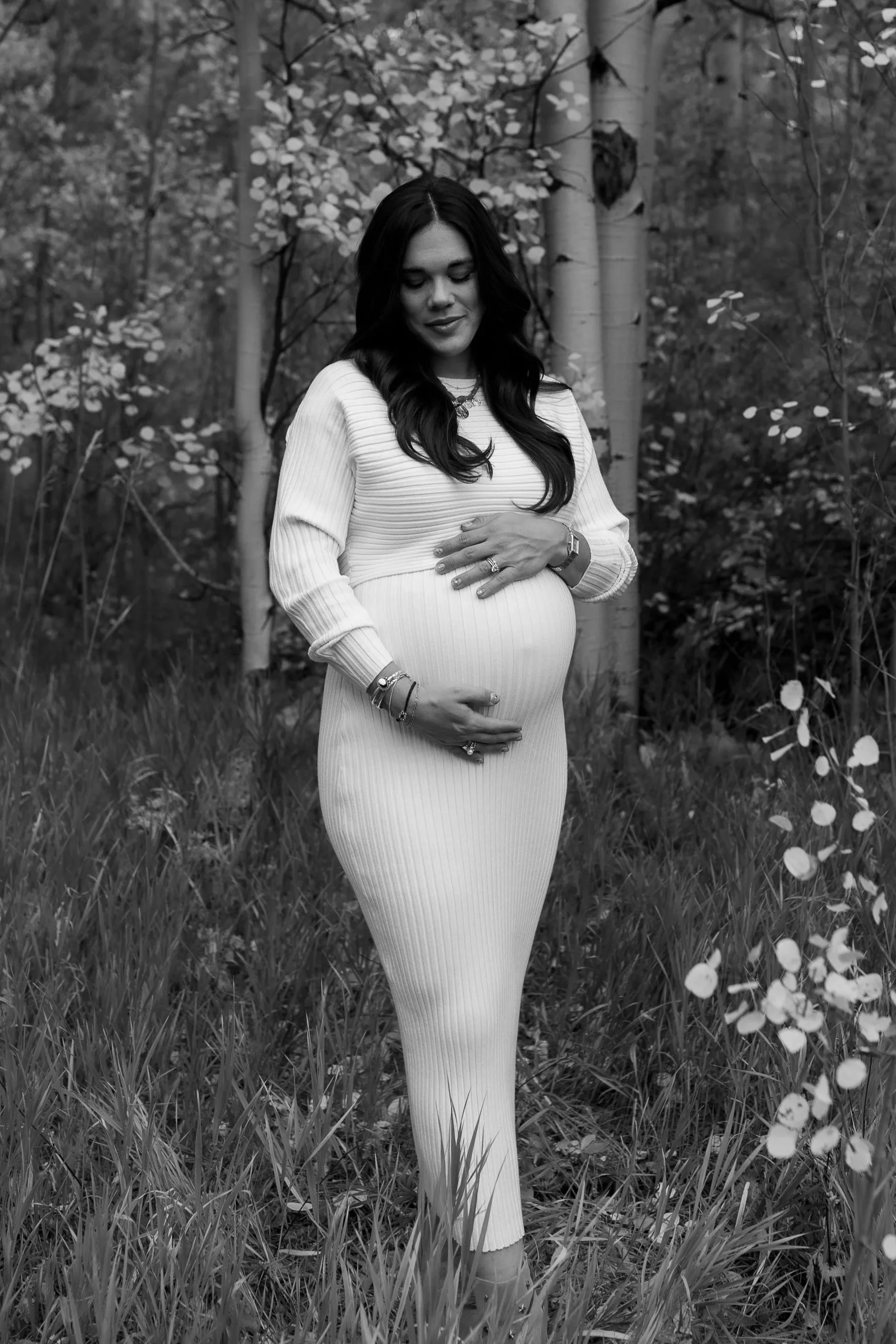 A pregnant woman standing in a forest setting, wearing a long, ribbed dress and looking down with her hands gently resting on her belly.