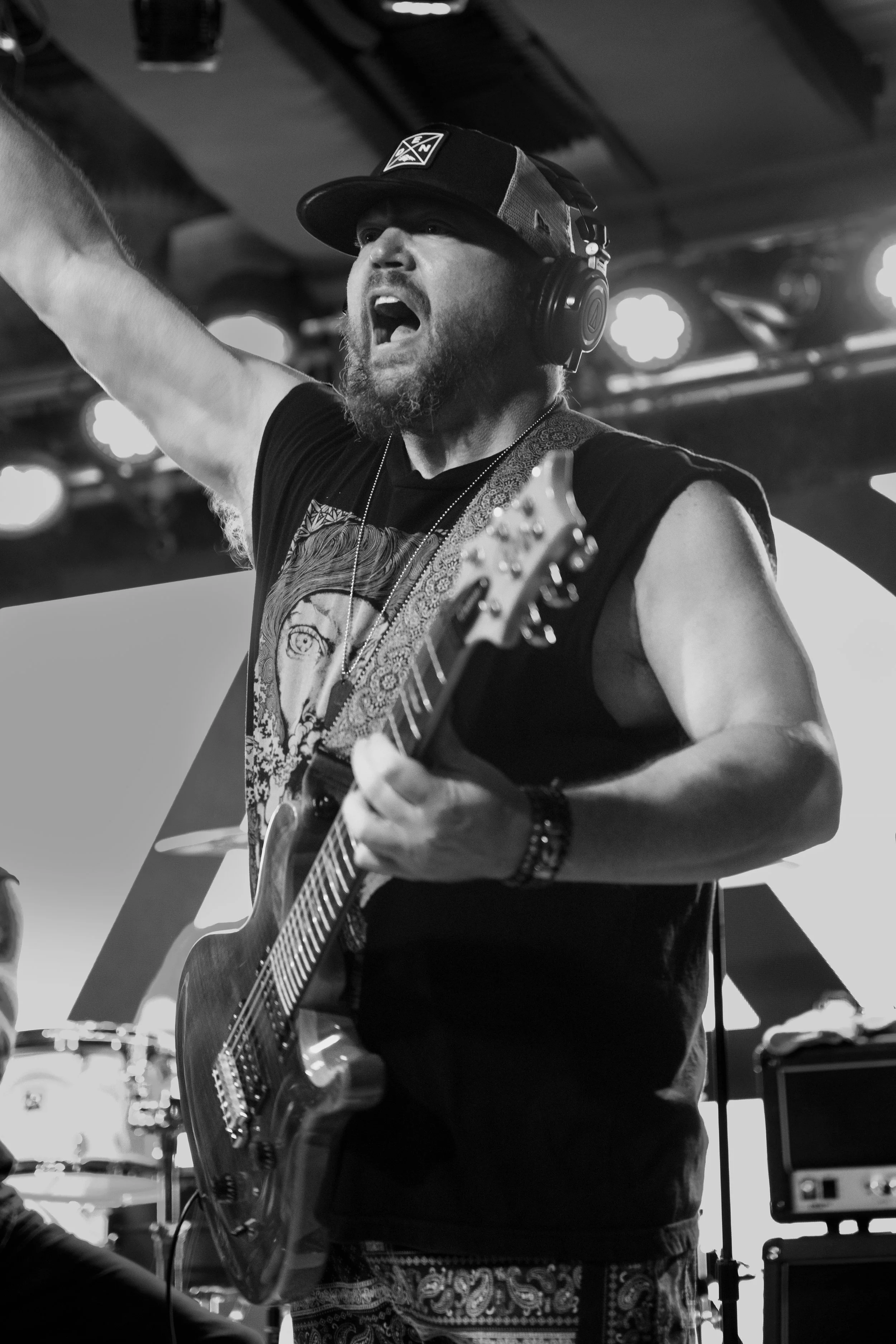 Guitarist of Shotgun Opera wearing a cap and headphones passionately playing an acoustic guitar on stage, with stage lights and musical equipment visible in the background.