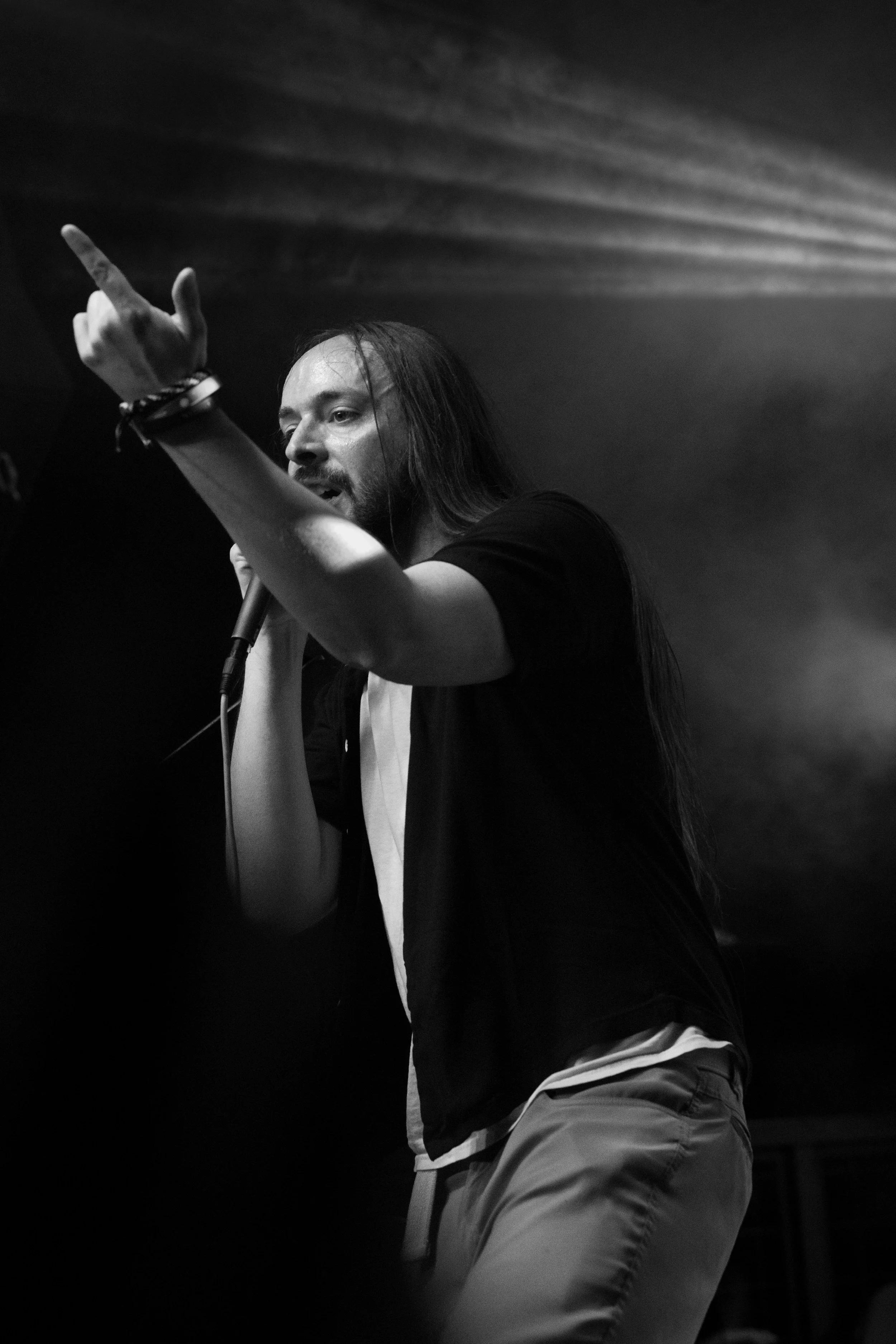 Front man of Shotgun Opera with long hair, wearing a black shirt and camouflage pants, is holding a microphone and raising his hand with his middle finger extended.