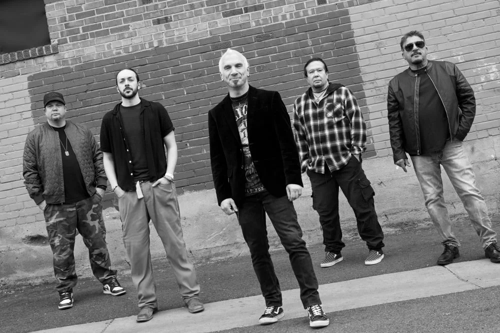 Five men standing in front of a brick wall, posing for a photo in casual and streetwear clothing, black and white photo.