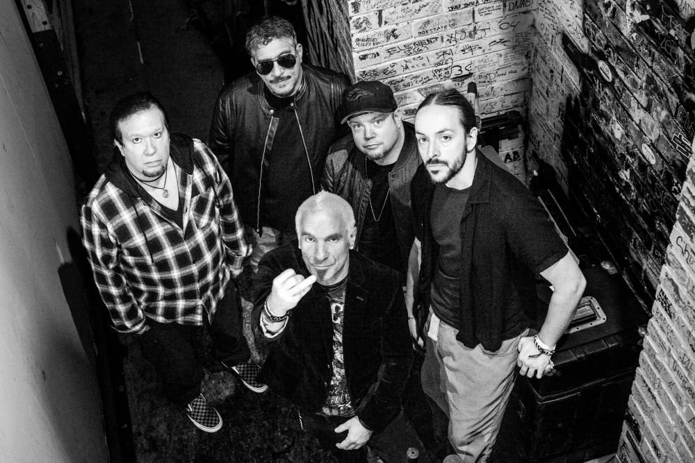 Five men posing in a narrow, graffiti-covered alley. One man is throwing a rock sign. The photo is black and white.