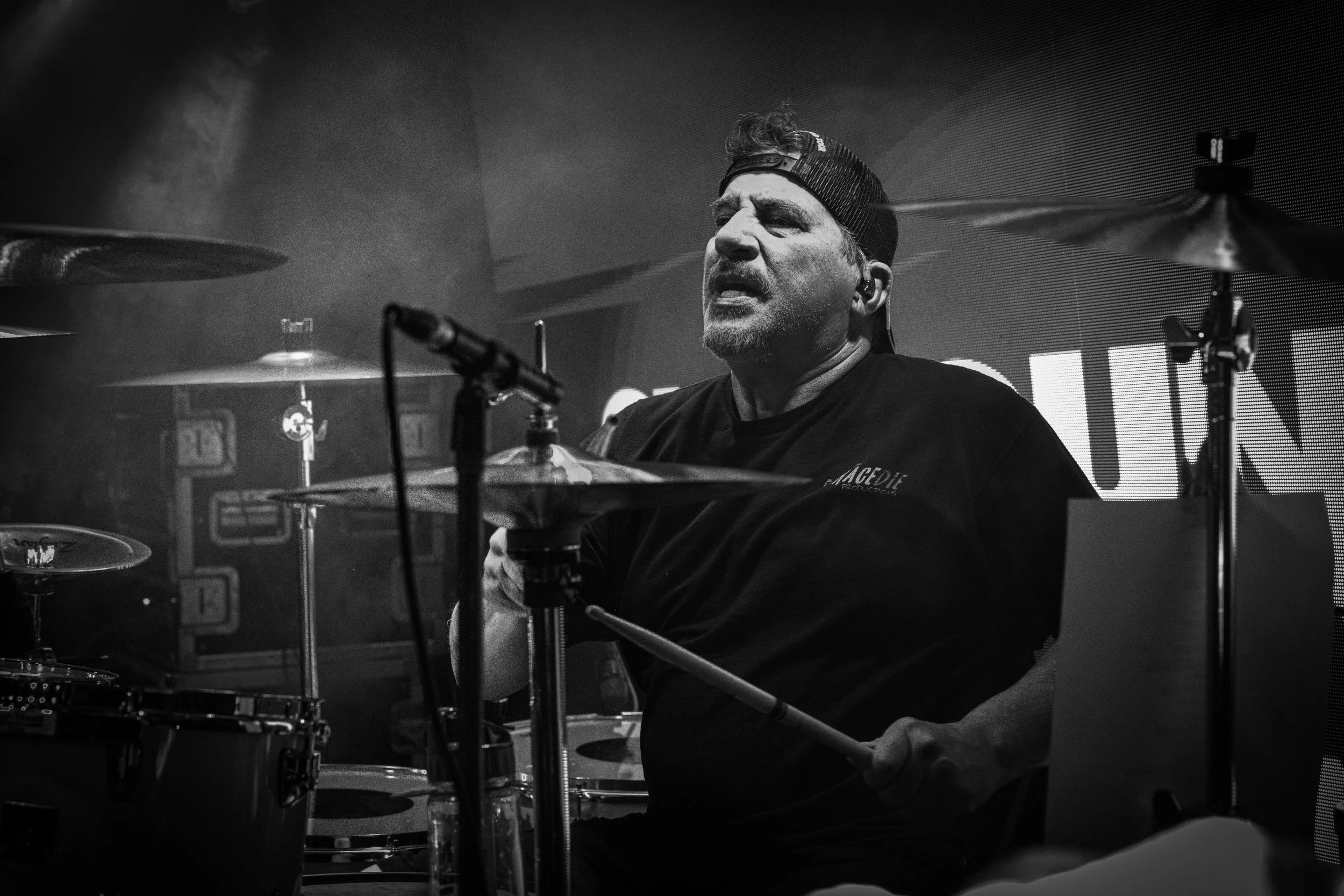 Drummer of Shotgun Opera with a beard and backwards cap playing drums during a live music performance, in black and white.