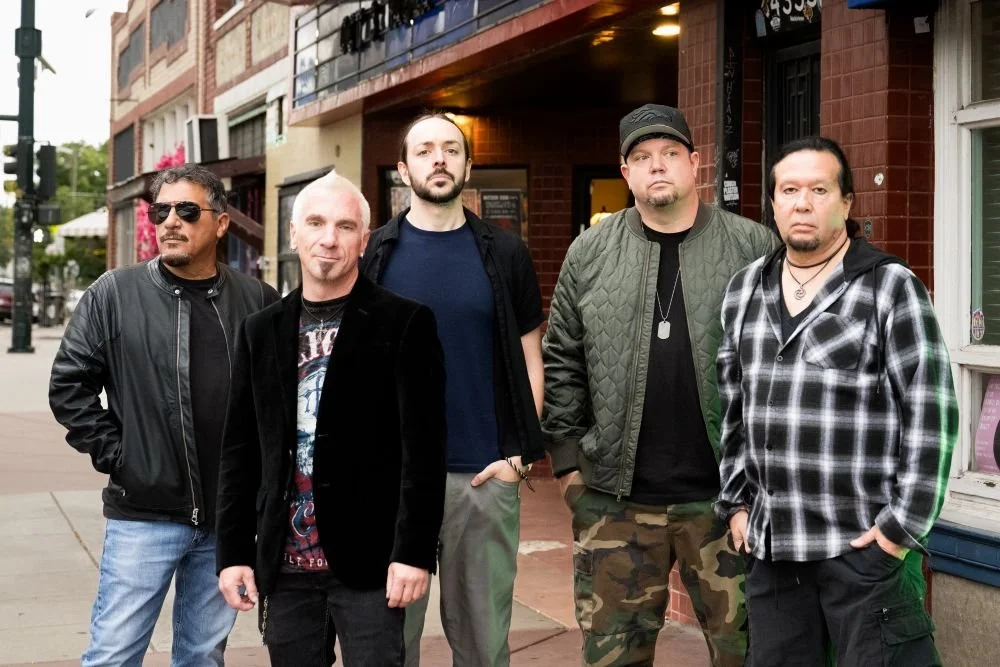 Five men standing on a city sidewalk in front of a brick building, posing for a photo, dressed in casual and punk style clothing.