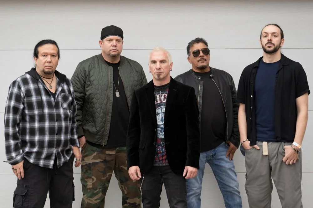 A group of five men standing against a gray wall, dressed in casual and edgy clothing, with serious expressions.