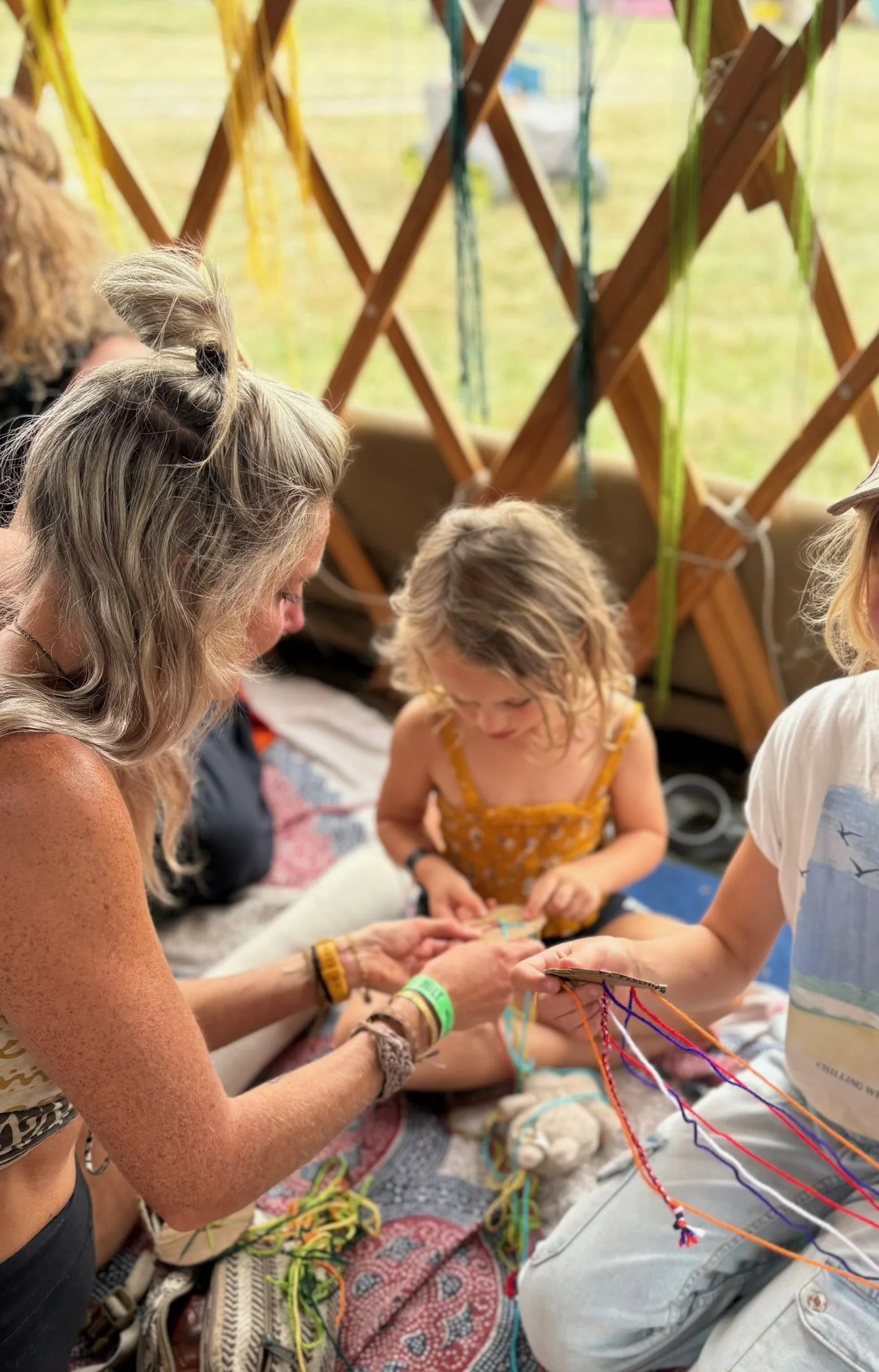 Group of people sitting on a blanket, engaged in a craft activity involving yarn and fabric, with a wooden lattice and greenery in the background.