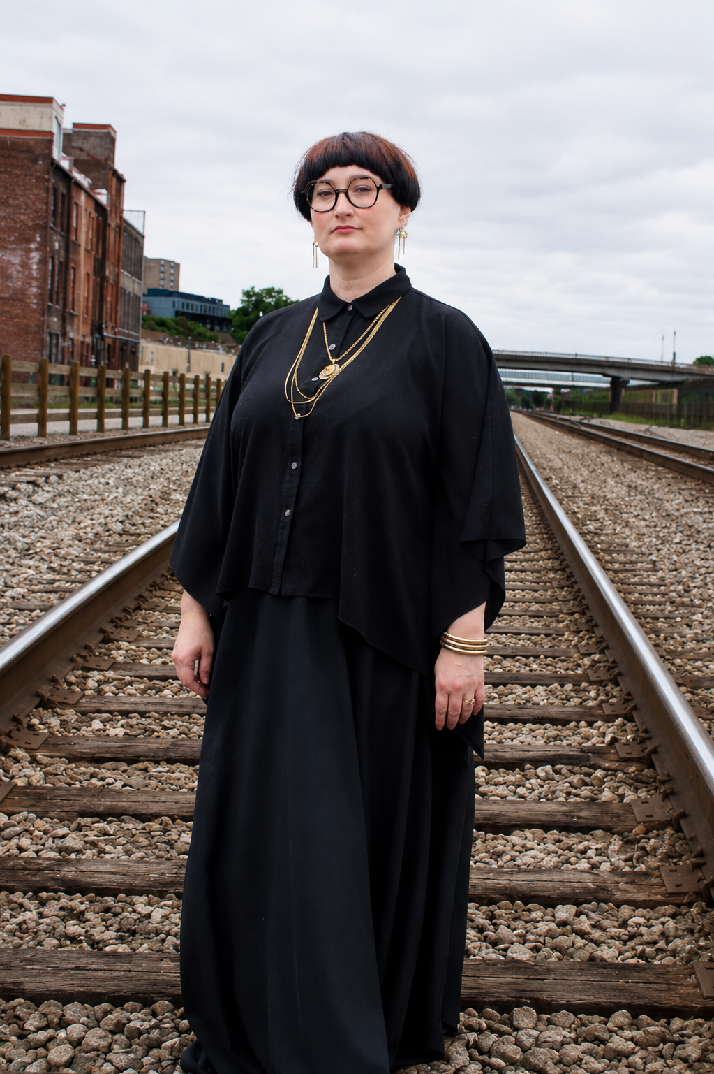 A woman with short brown hair wearing glasses, a black lace top with sheer floral details, and long geometric earrings, standing in front of a brick wall with framed pictures.