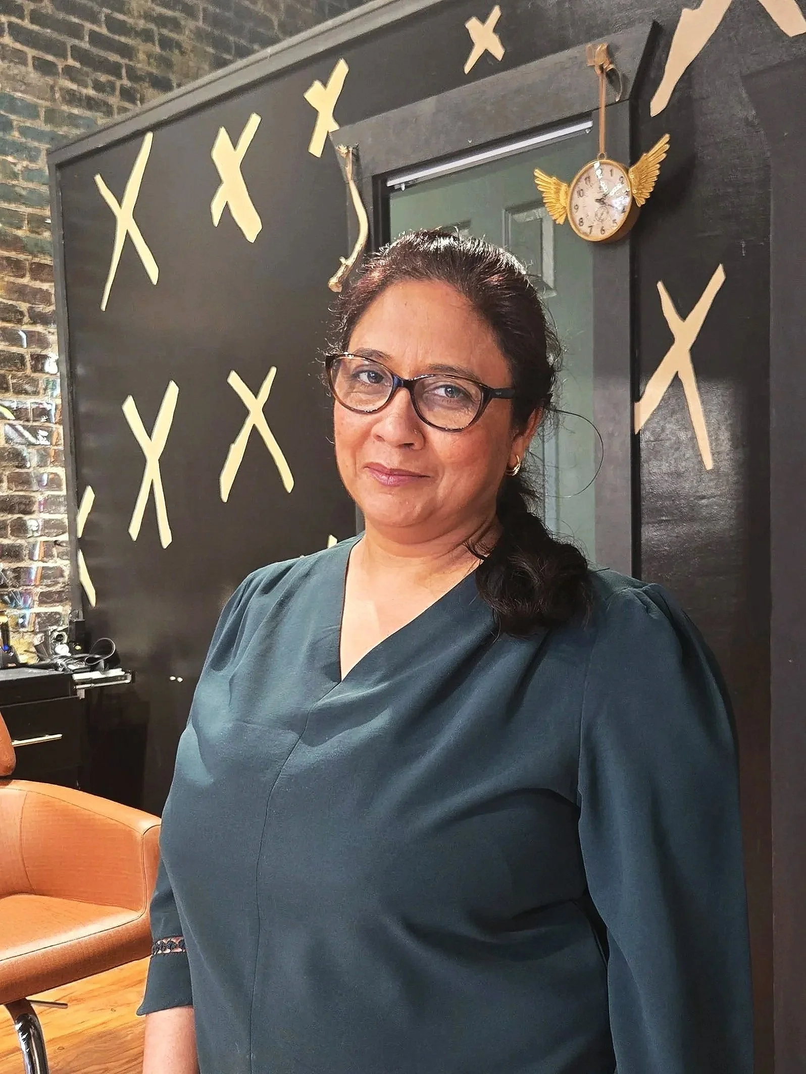 A woman with glasses stands in front of a black wall decorated with yellow Xs and a clock with wings.