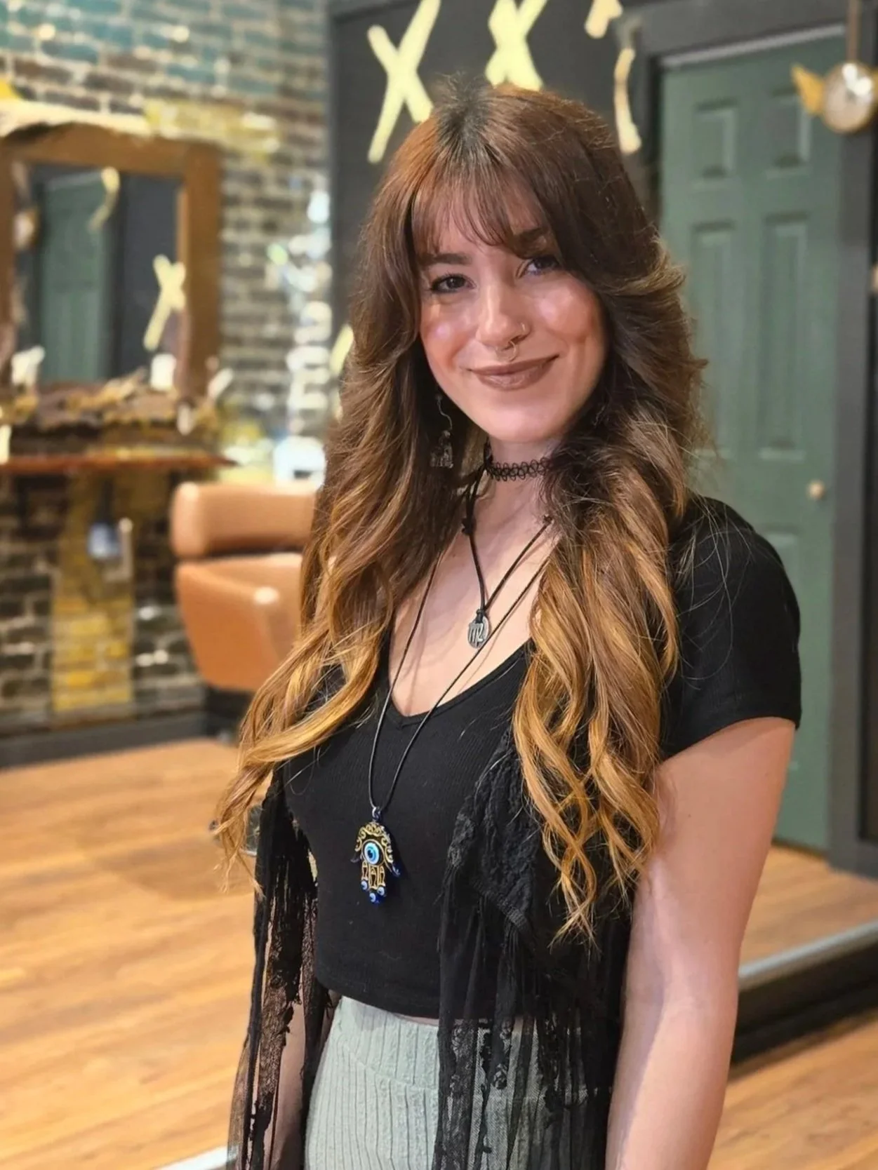 A young woman with long, wavy, ombre hair and light skin, smiling, wearing layered necklaces, a black choker, and a black top with a sheer black lace overlay. She is standing inside a room with a decorated wall background.