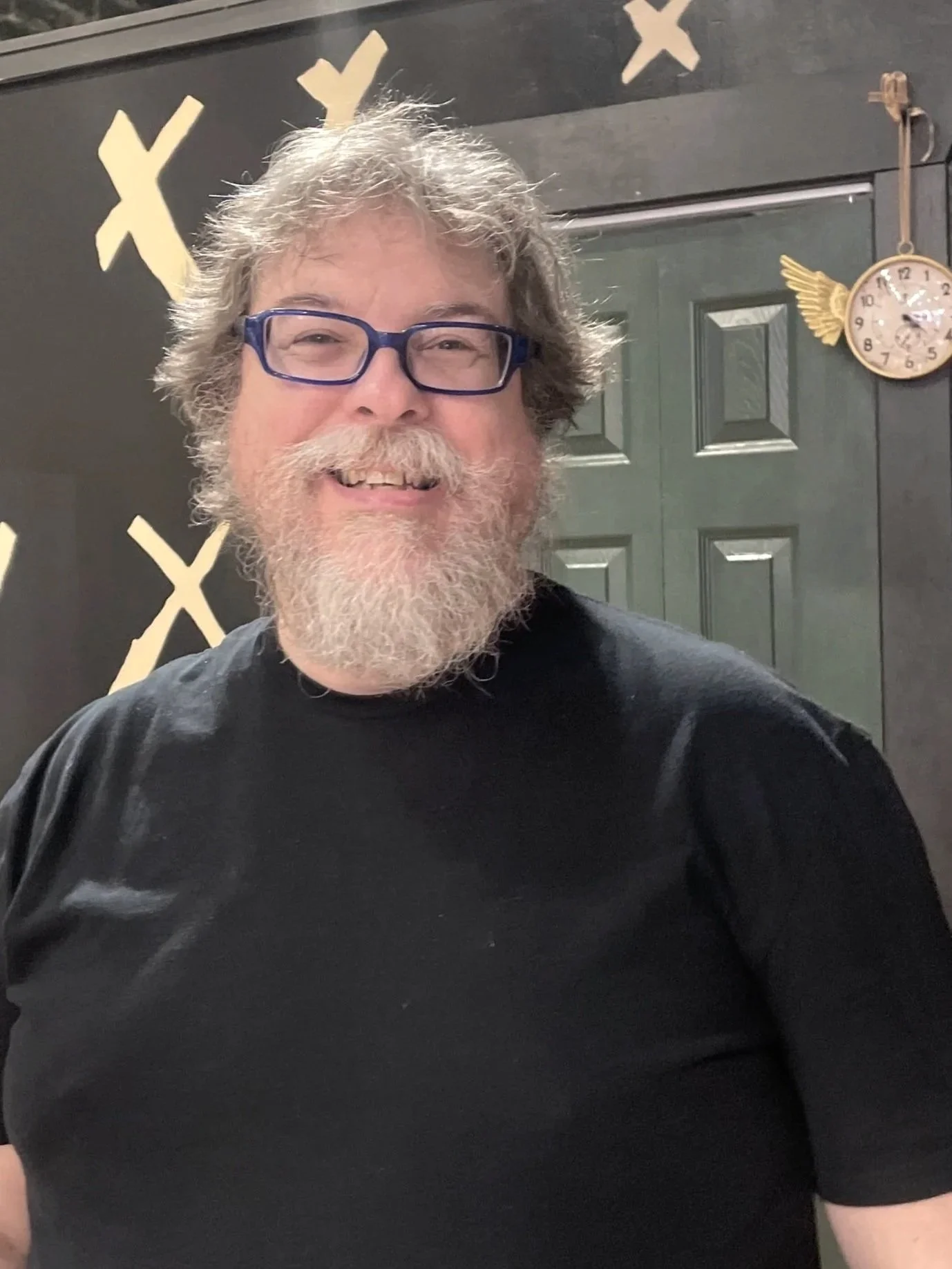 A smiling man with gray hair, glasses, and a beard wearing a black t-shirt. Behind him is a dark wall with yellow X patterns, a green door, a clock with wing and circle design, and some hanging decorations.