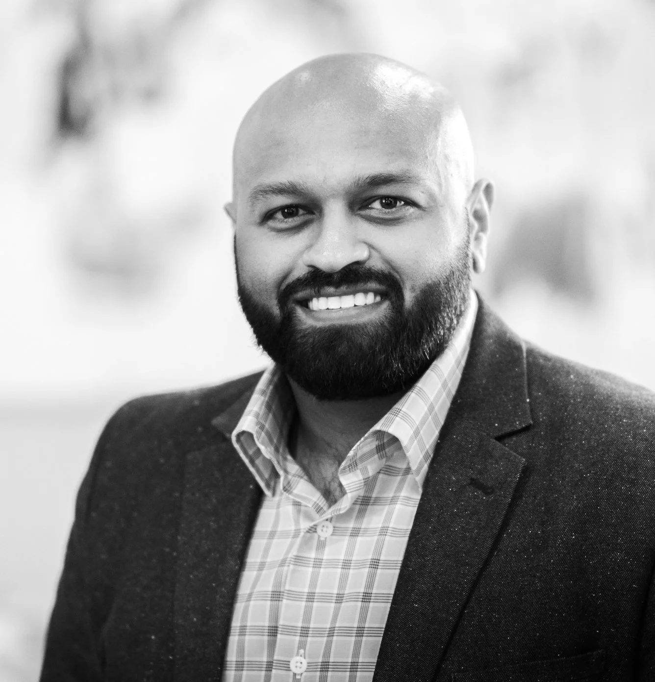 A smiling man with a beard and shaved head, wearing a suit jacket and checkered shirt, looking at the camera.