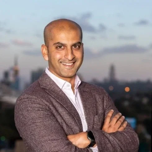 A man in a blazer smiling with arms crossed against a city skyline at dusk.
