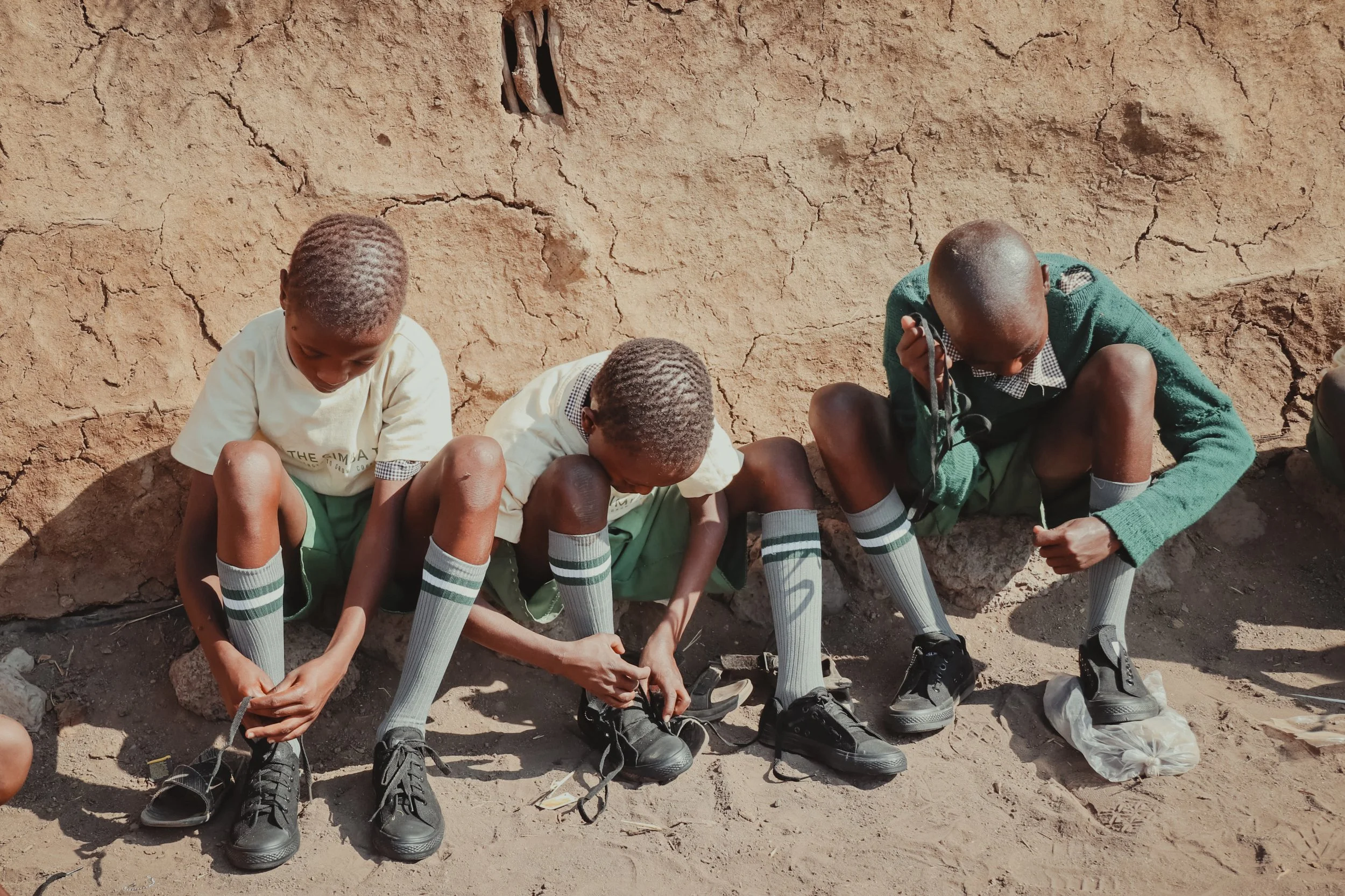 Three schoolboys sitting on the ground against a cracked earthen wall, fastening their black shoes, wearing school uniforms with white shirts, green skirts or shorts, and knee-high socks.