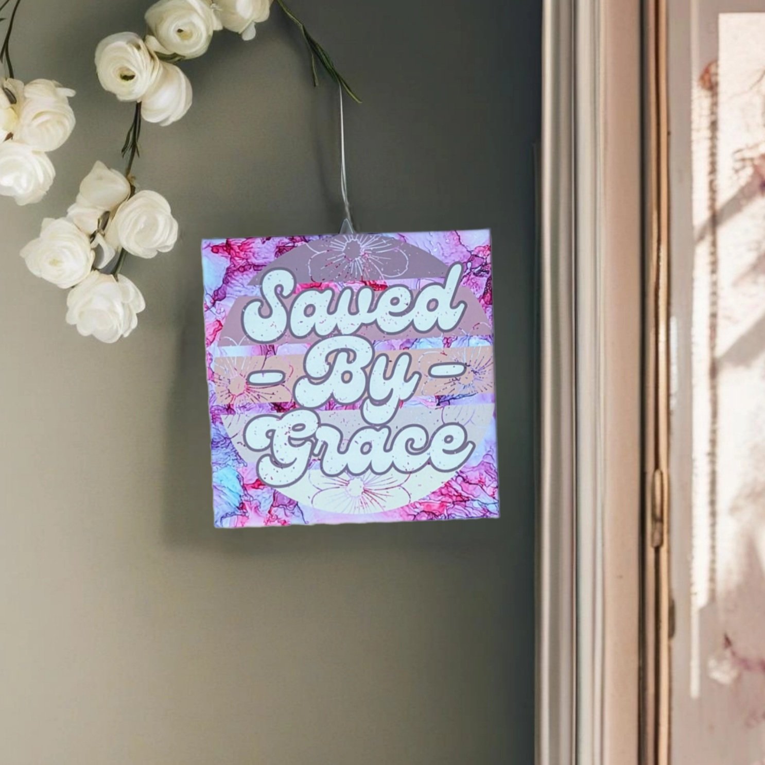 8 x 8-inch ceramic tile with pinkish alcohol ink background and the words "Saved By Grace" positioned on a wall near a window. There are also flowers on the opposite side of the tile