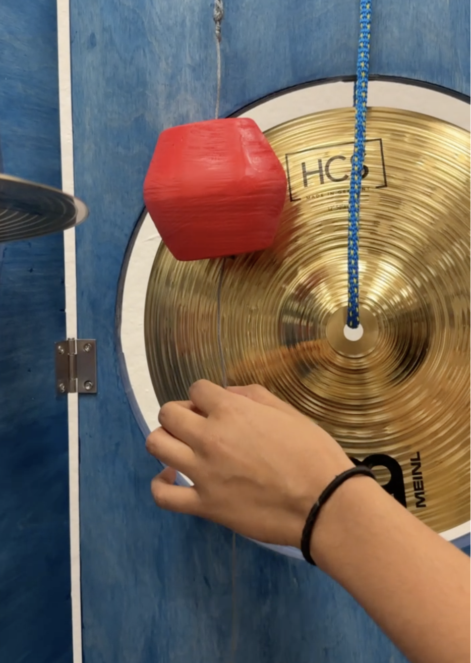 A person's hand holding a red mallet close to a brass cymbal, with a blue and yellow striped rope hanging above it and a blue wall in the background.