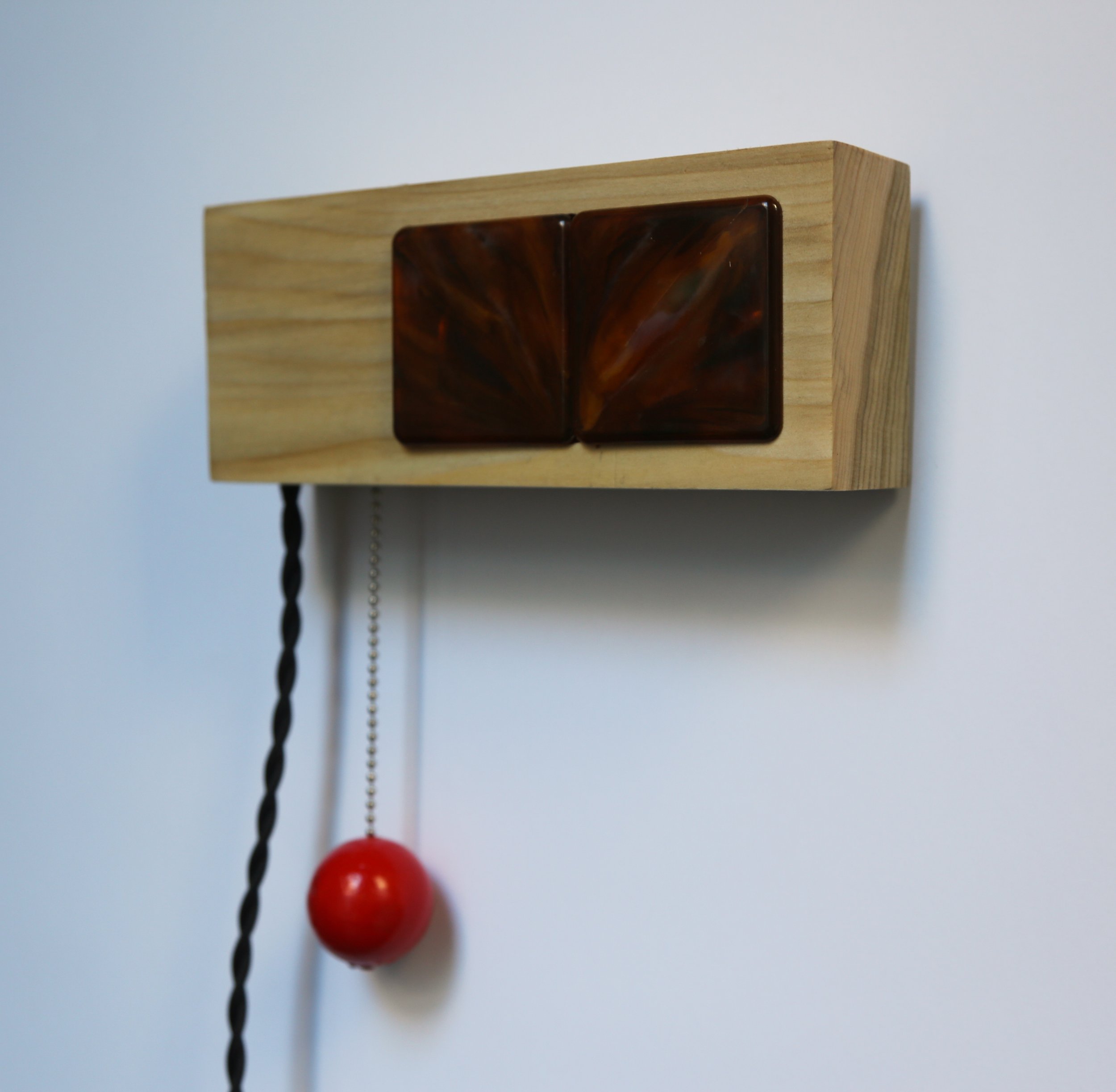A wall-mounted wooden box with two brown toggles, a black twisted pull cord with a red ball at the end, and a white chain pull.