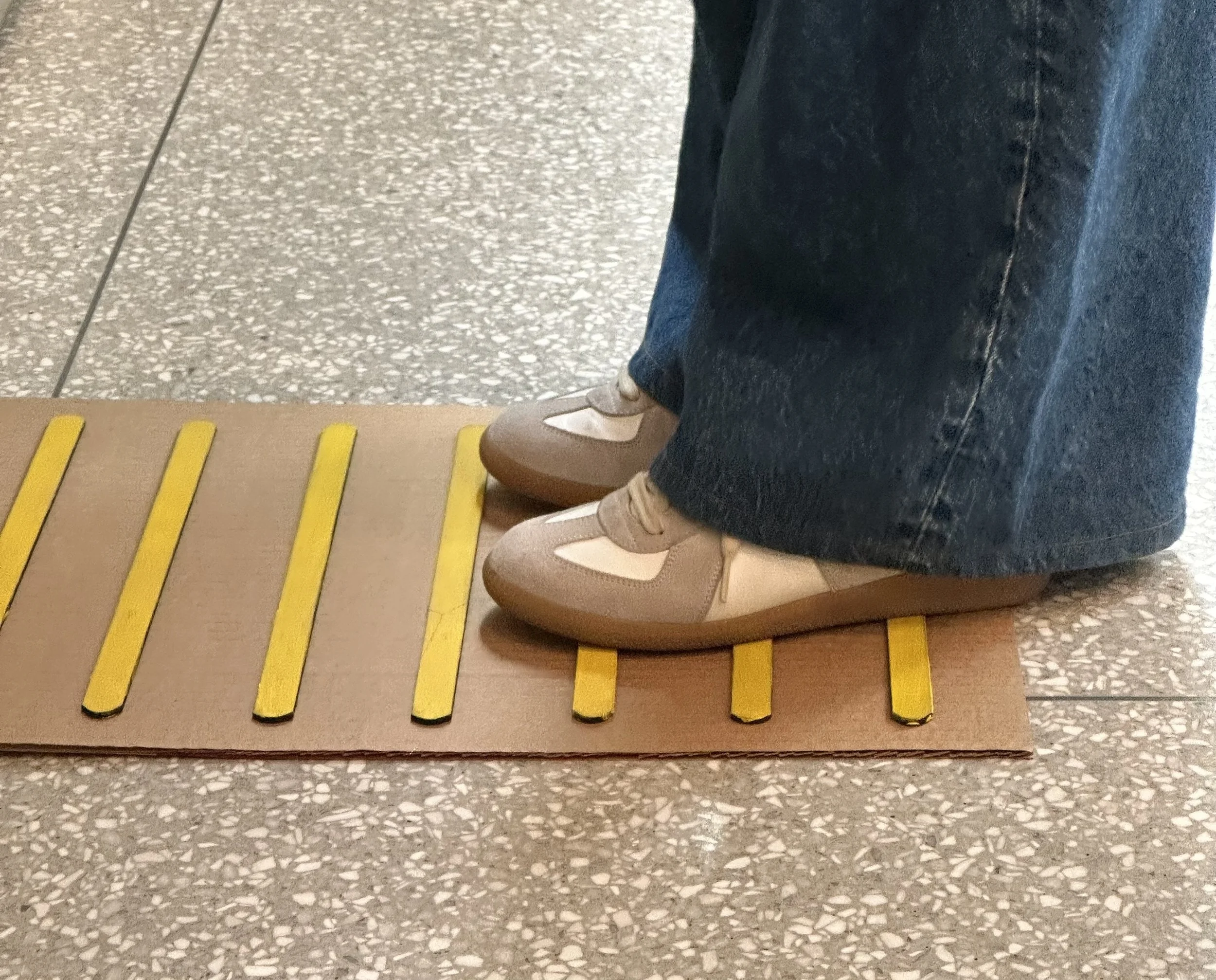 Person standing on a tactile warning strip with yellow and black lines, wearing blue jeans and beige shoes, on a speckled gray floor.