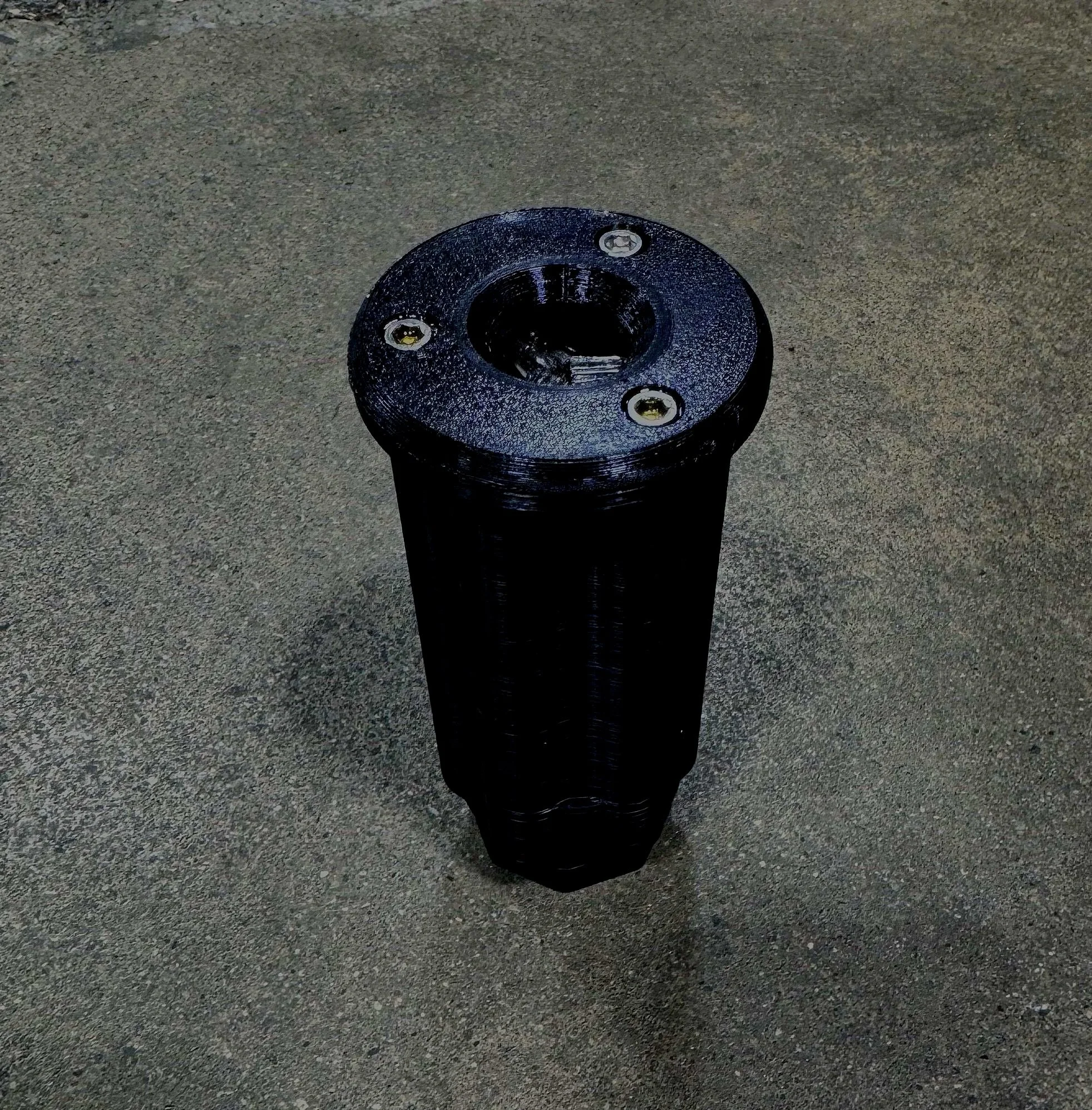 A black cylindrical object with a round top and three screws on the top surface, placed on a gray textured surface.