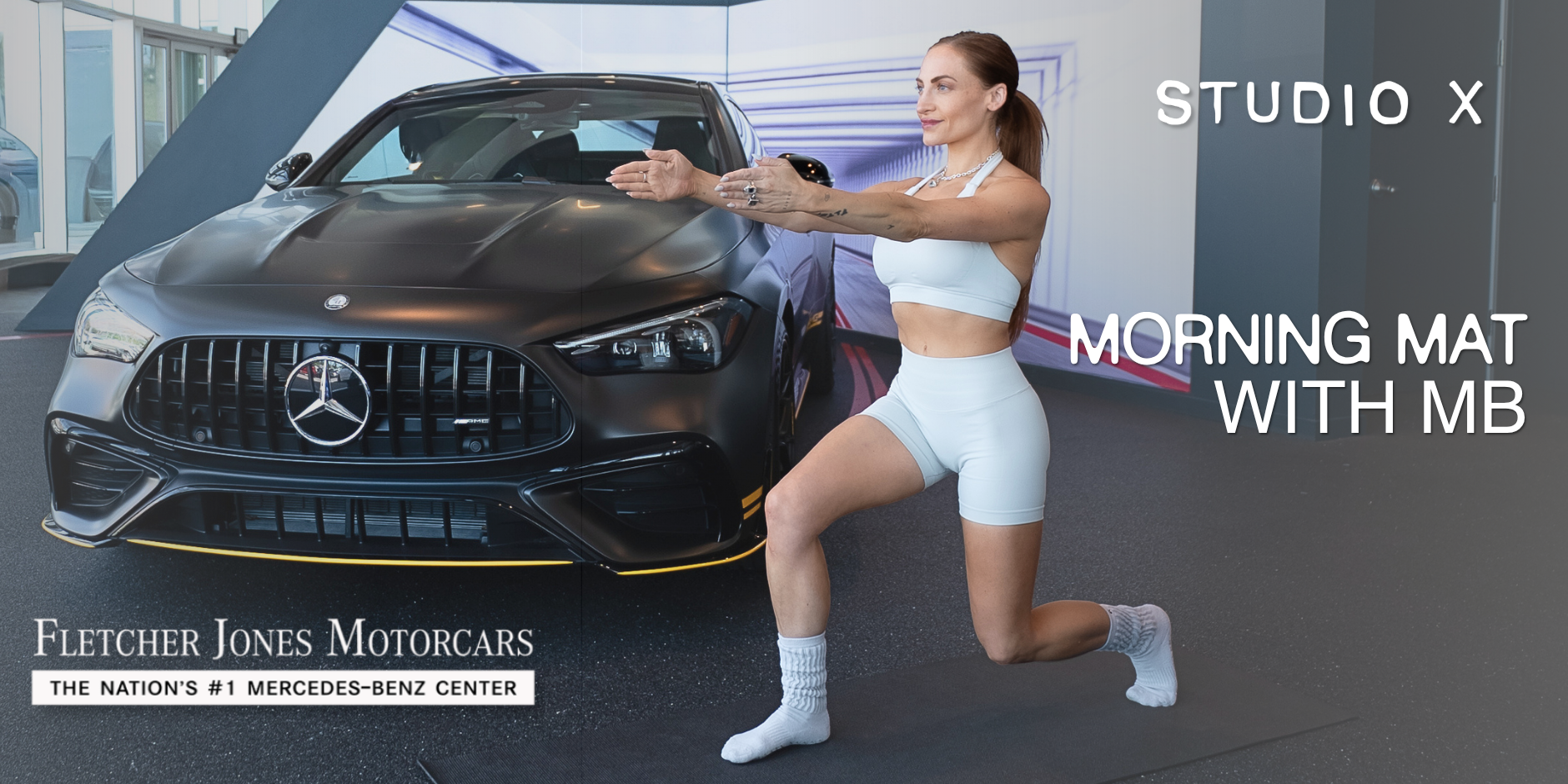 A woman in white workout clothes is doing a fitness pose kneeling on one knee in front of a black Mercedes-Benz car in a showroom with a digital display behind her. The text on the image reads 'STUDIO X Morning Mat with MB' and 'Fletcher Jones Motorcars, the nation's #1 Mercedes-Benz center.'