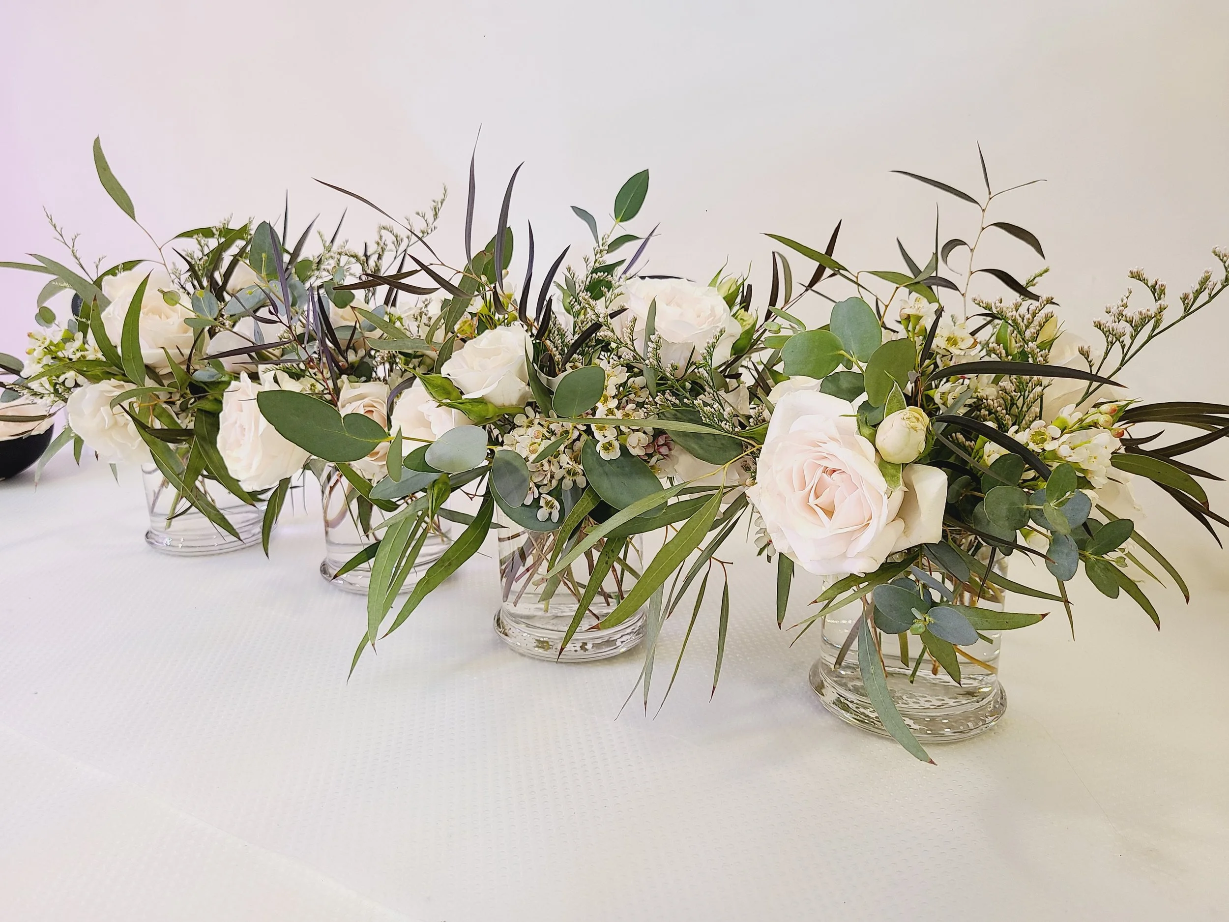 Five glass vases with white roses and greenery on a white table.