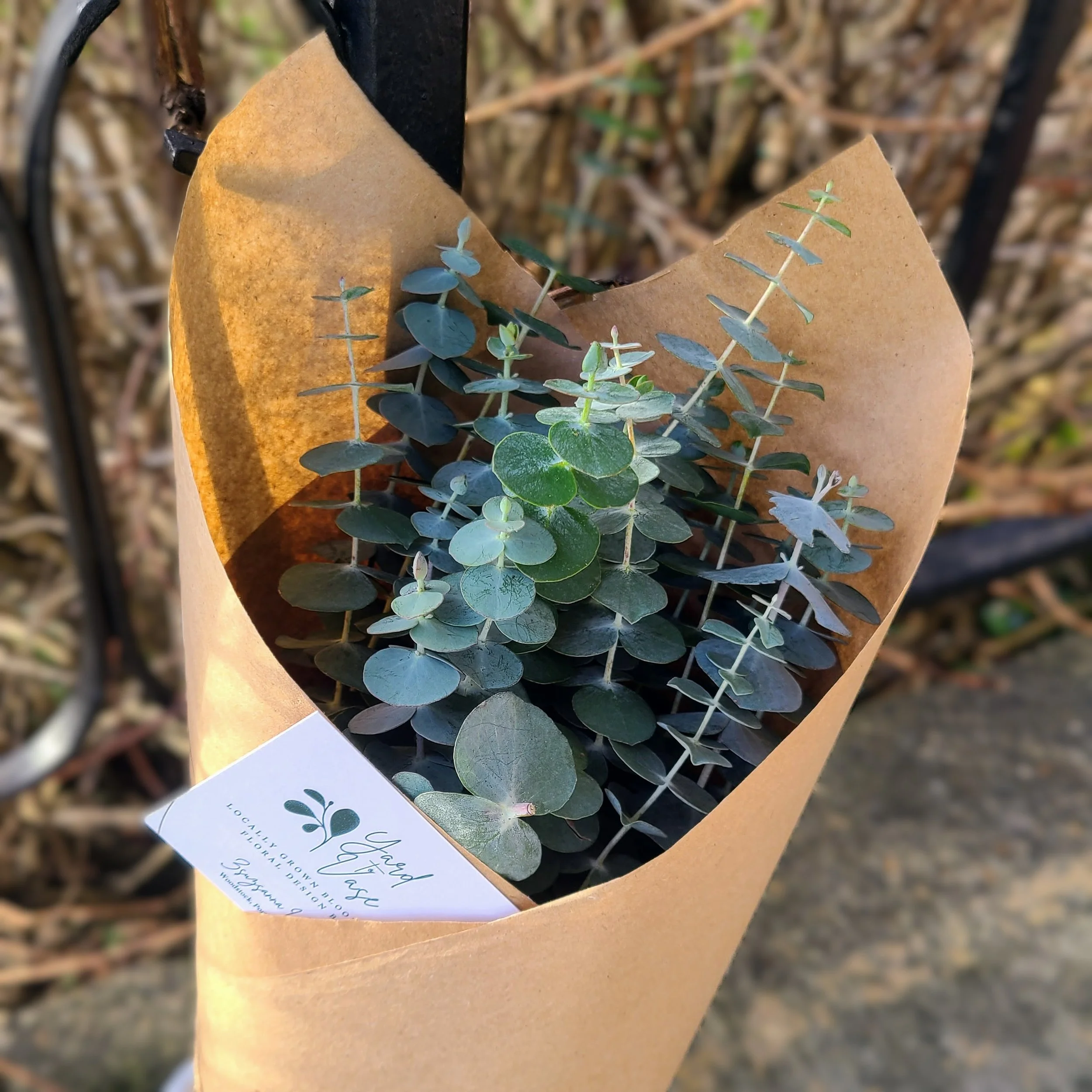 Bouquet of eucalyptus leaves wrapped in brown paper with a gift card attached, outdoors.