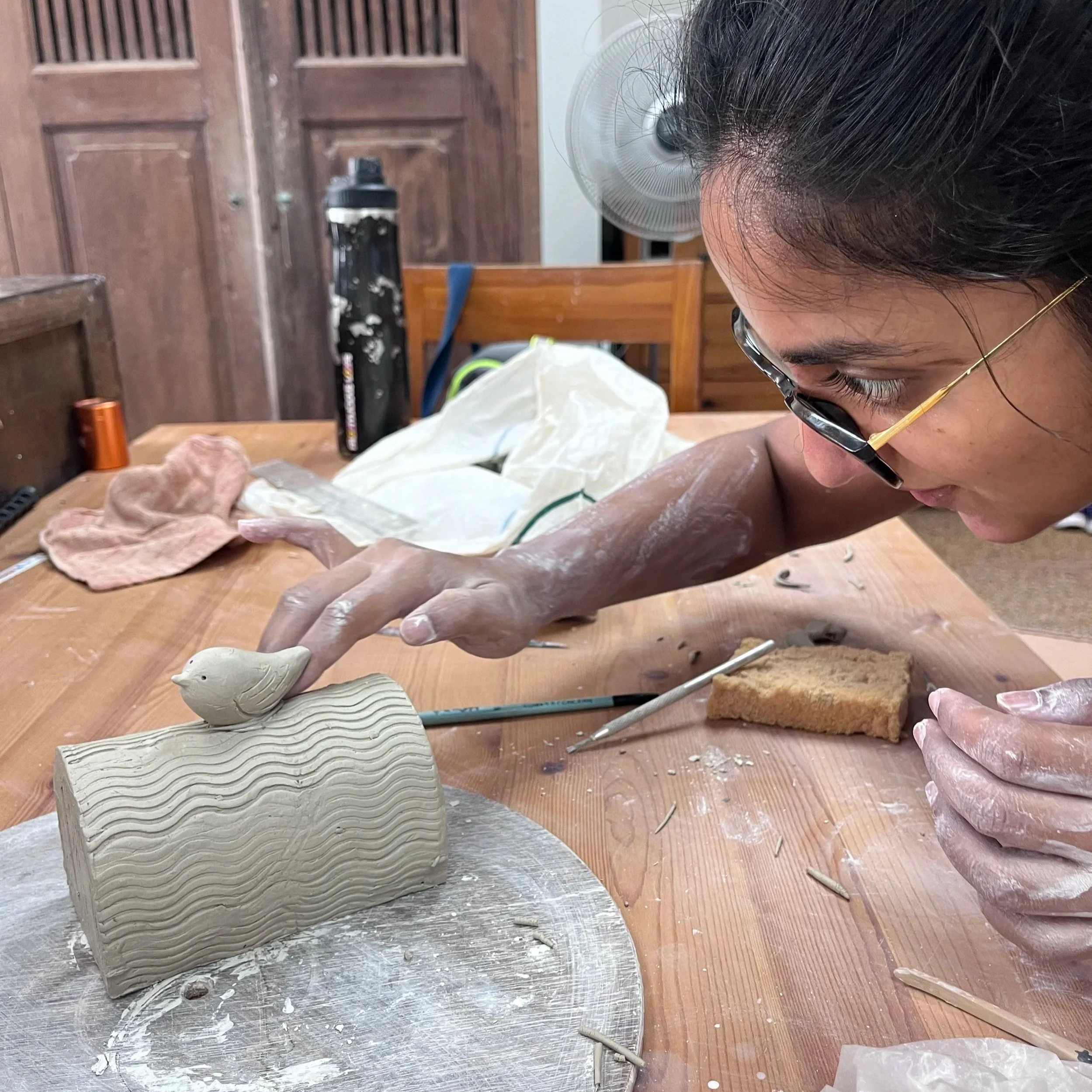 A woman leans over a wooden workbench, carefully adjusting a small clay bird perched atop a textured, wavy-patterned ceramic cylinder. Her hands are dusted with clay, and various pottery tools and a sponge sit on the table around her.