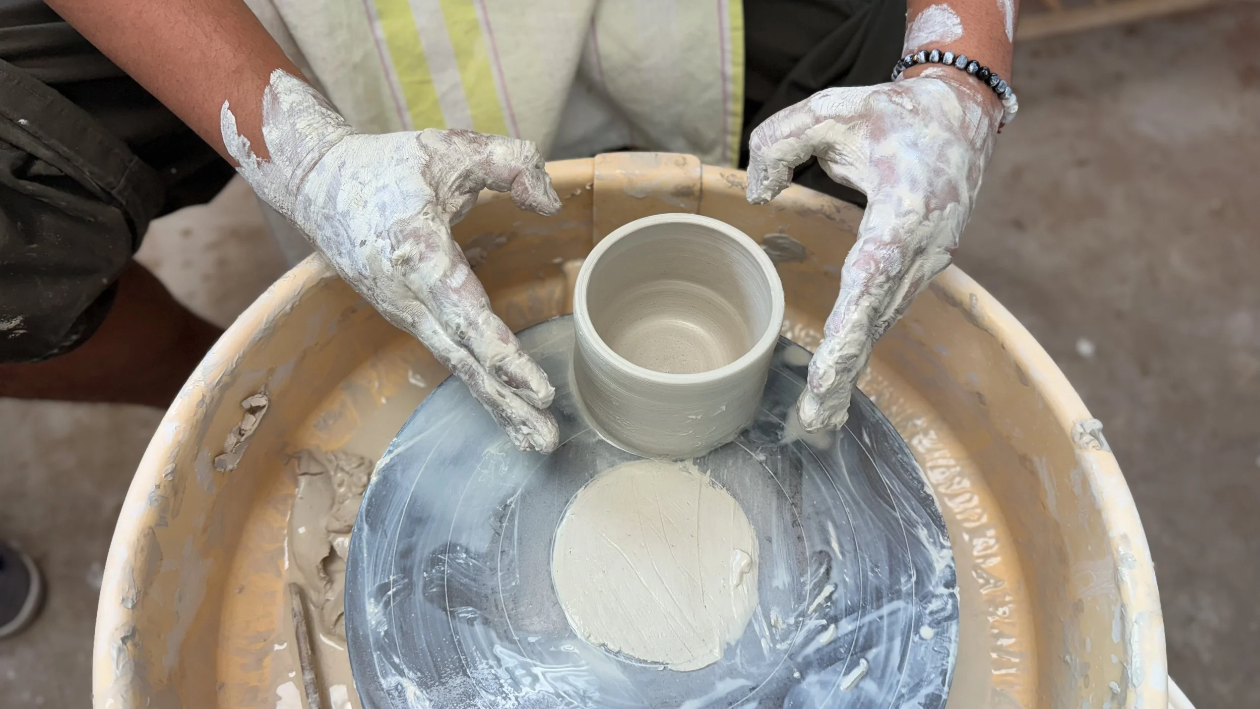 A close-up, overhead view of a potter's hands, heavily coated in wet clay, positioned around a small cylindrical pot on a pottery wheel.