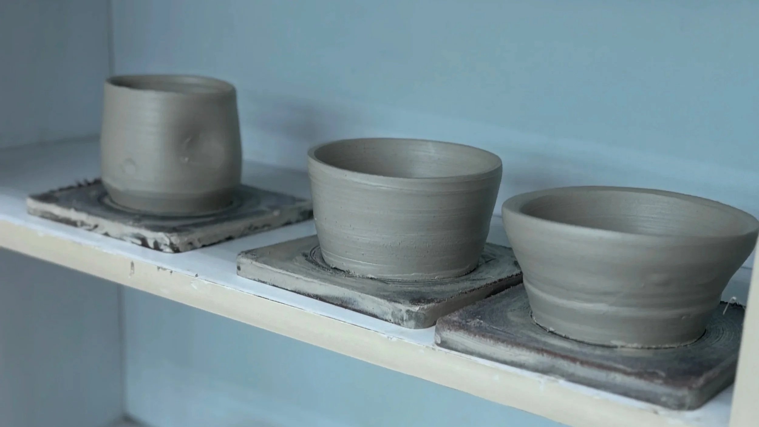 Three hand-thrown gray clay pots in various shapes sit on square wooden bats along a clean, minimalist white shelf.