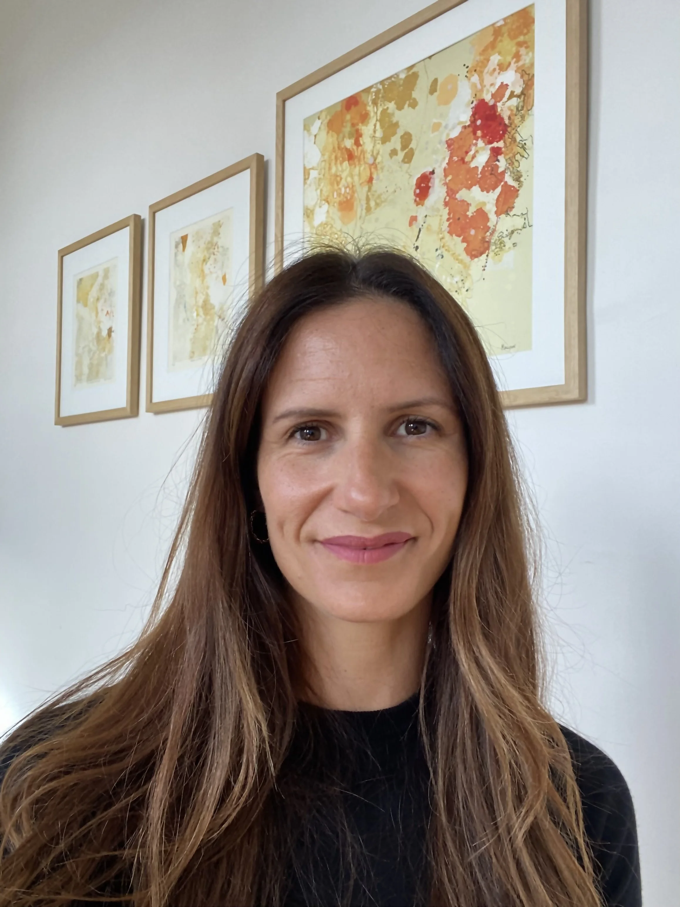 Sonia Dempsey in her consulting room at Stable Health in North Fitzroy. Sonia has long brown hair and a black top, smiling, in front of three framed watercolor art prints with shades of orange, yellow, and red on a white wall.