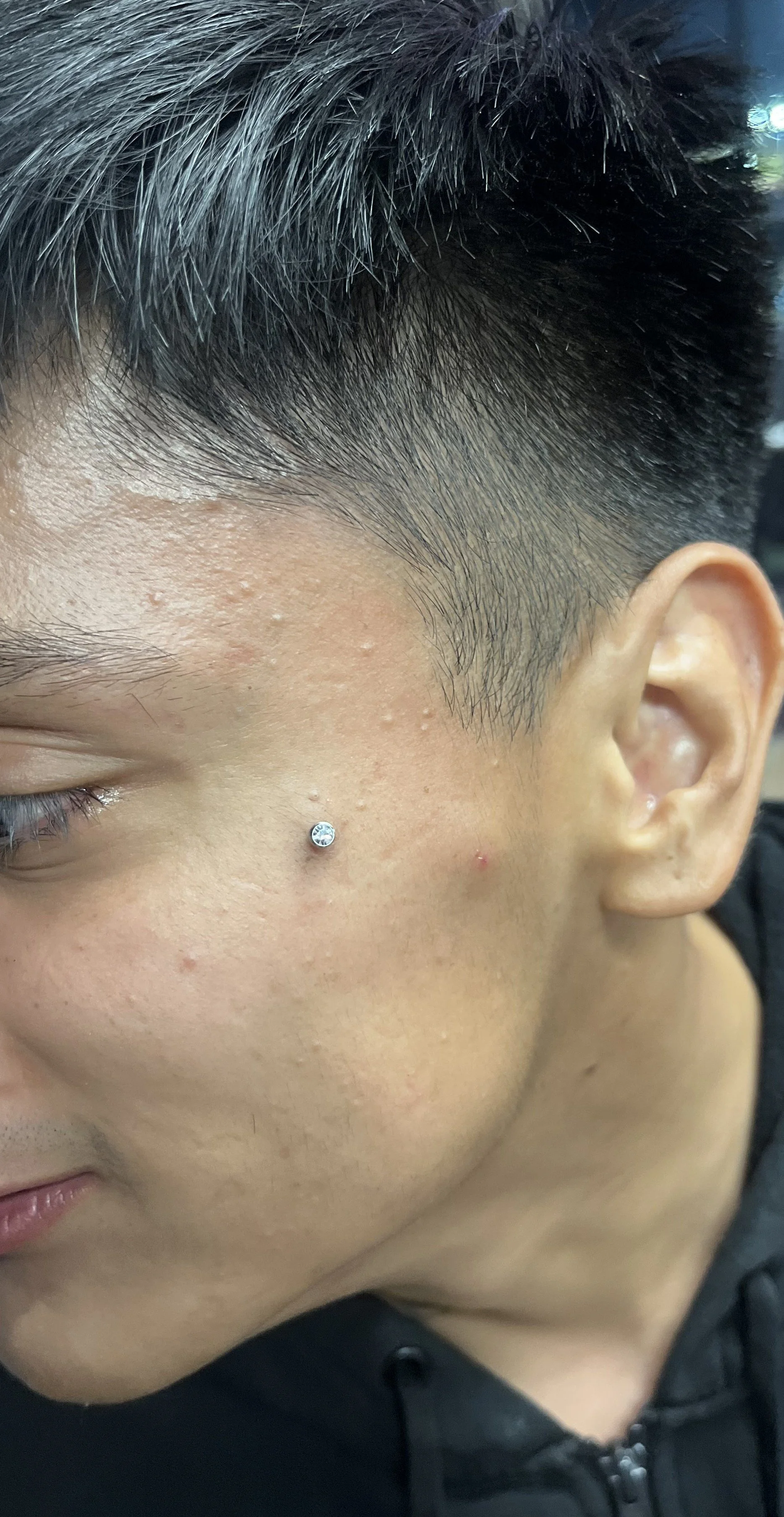 Close-up of a young person's face showing a silver nose stud, short dark hair, and lightly textured skin.