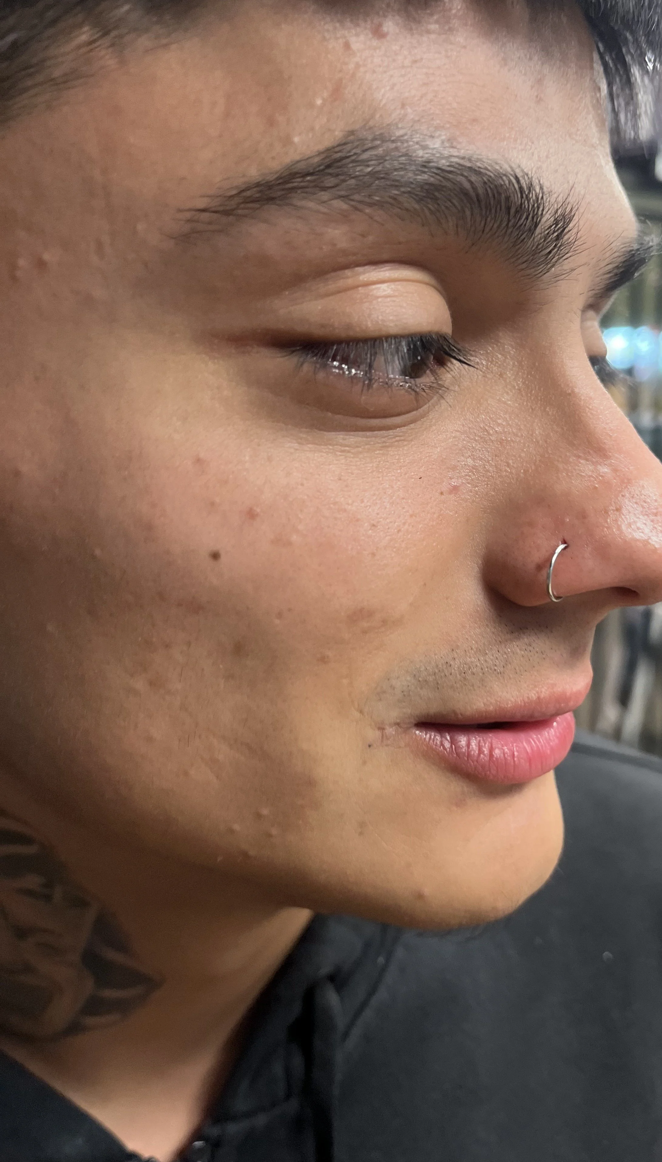 Close-up side profile of a person's face with a visible nose piercing, short dark hair, dark eyebrows, and textured skin.