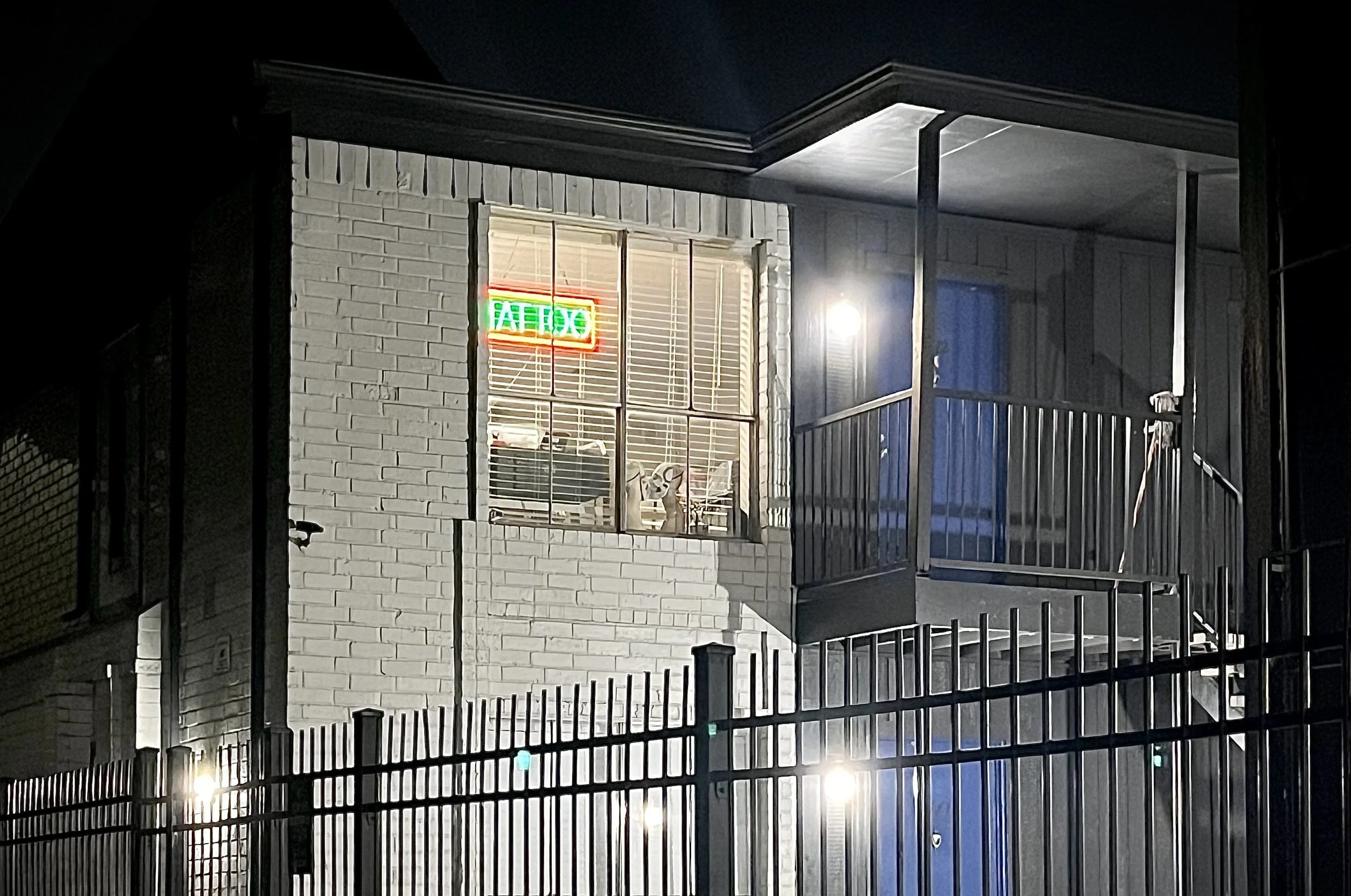 Night view of a two-story building with a white brick exterior, illuminated by exterior lights. A window on the second floor displays a 'TATTOO' neon sign and a dog looking out.