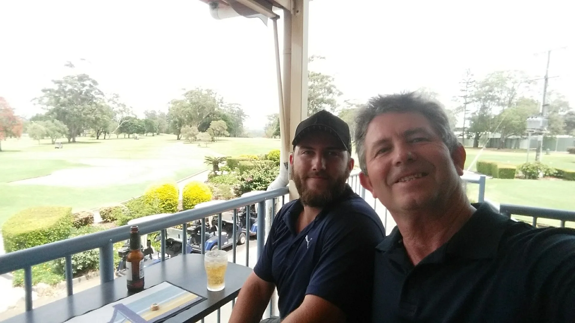 Two men sitting at an outdoor table on a balcony, with a golf course in the background; one man is smiling and taking a selfie, the other has a beard and is wearing a black cap.